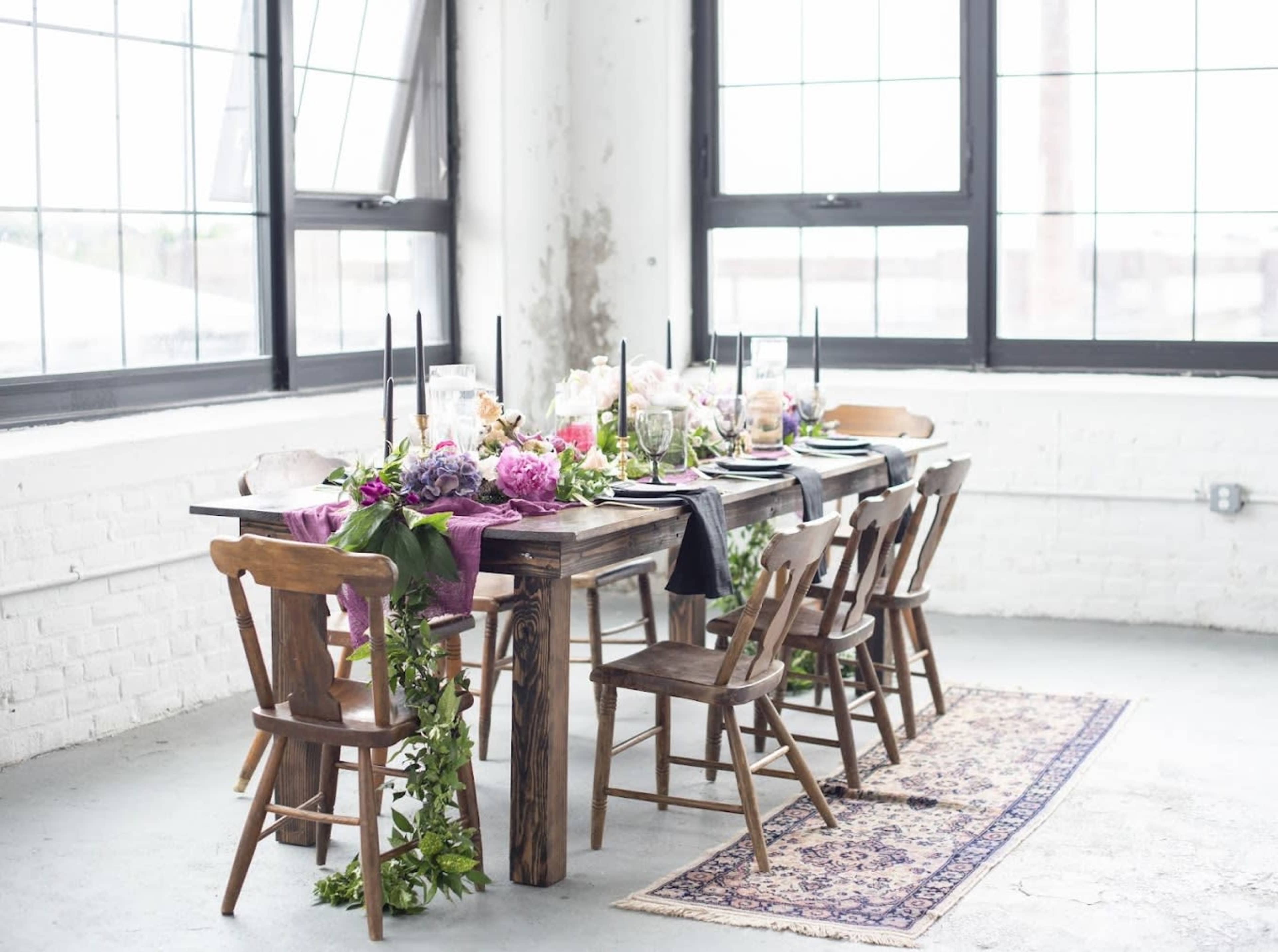 A long wooden dining table is set with colorful floral arrangements, candles, and tableware, surrounded by wooden chairs in a bright, airy room with large windows.