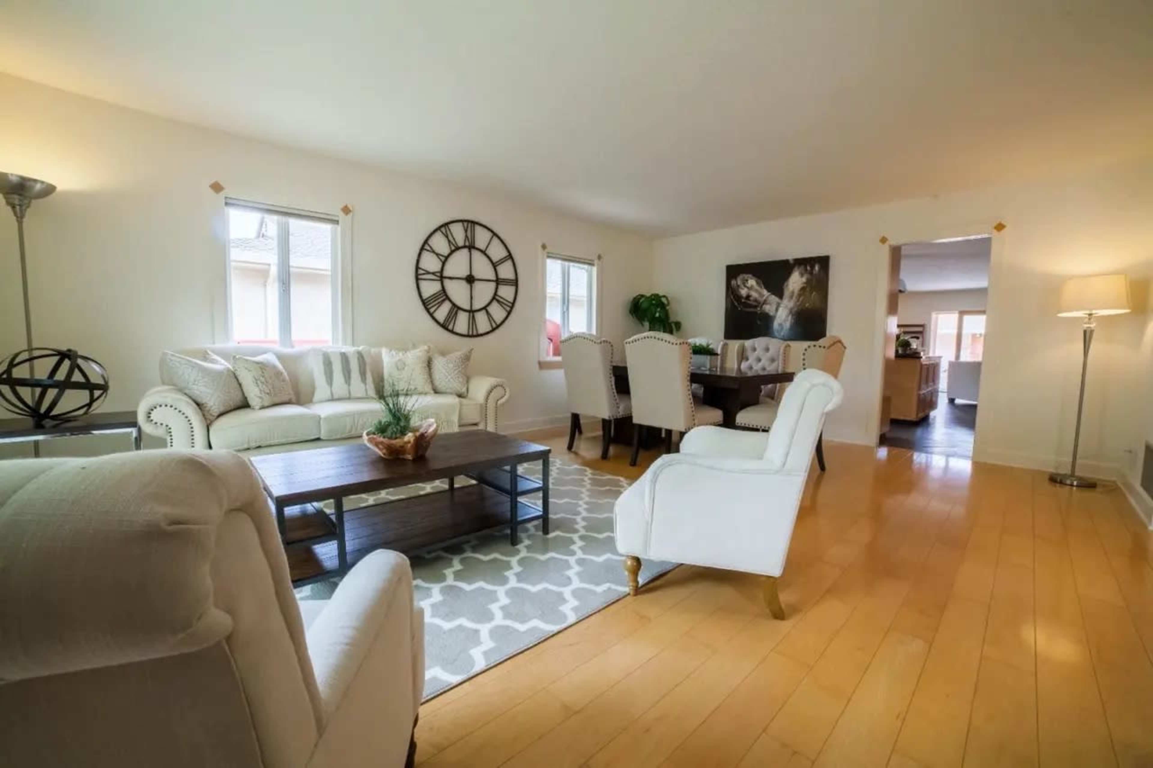 A bright living room with a white couch, wooden floors, and a large wall clock, leading into a dining area in the background.