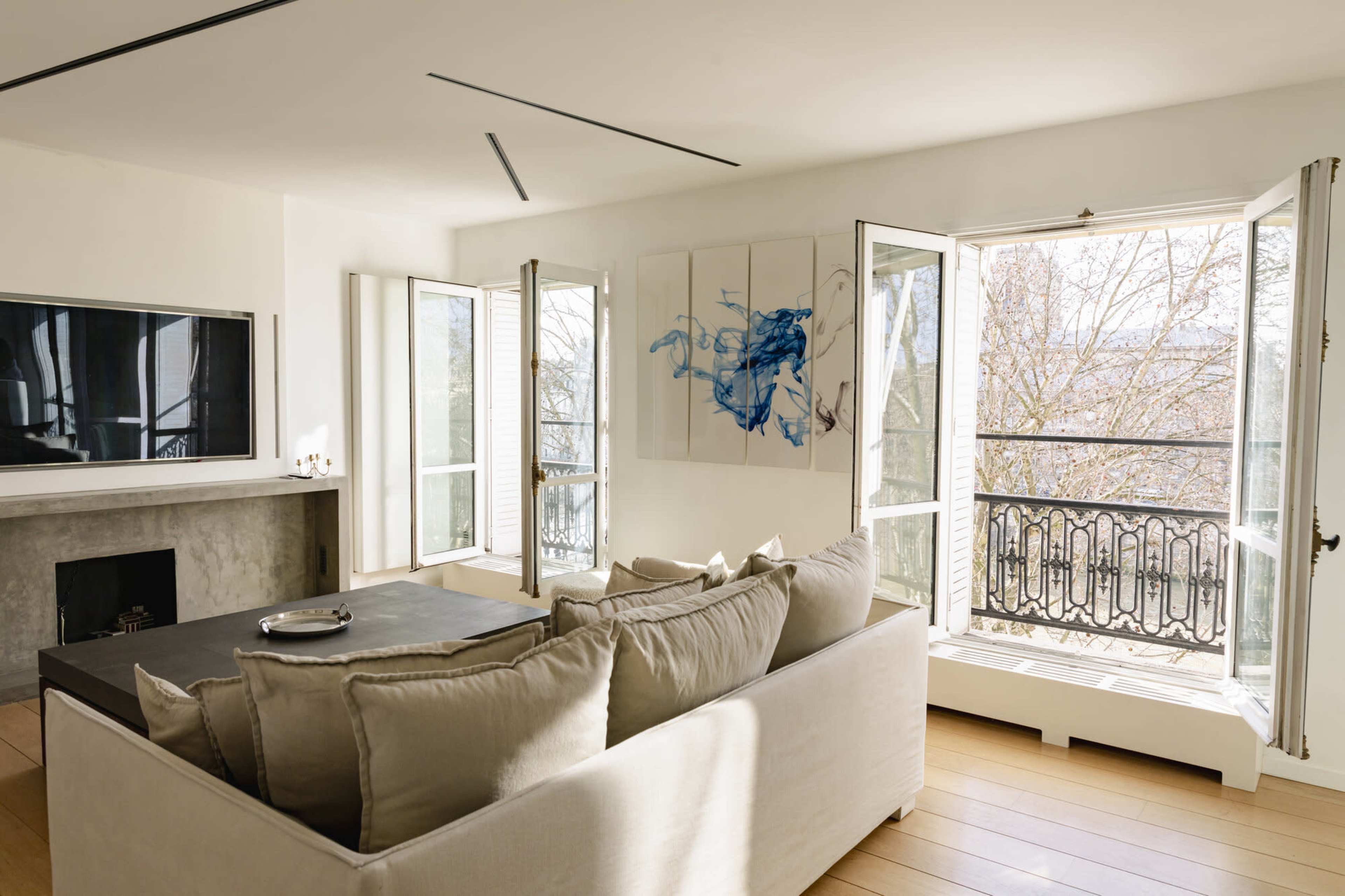 A modern living room features a light-colored sofa, a large flat-screen TV, and two open windows with views of a balcony and trees outside.