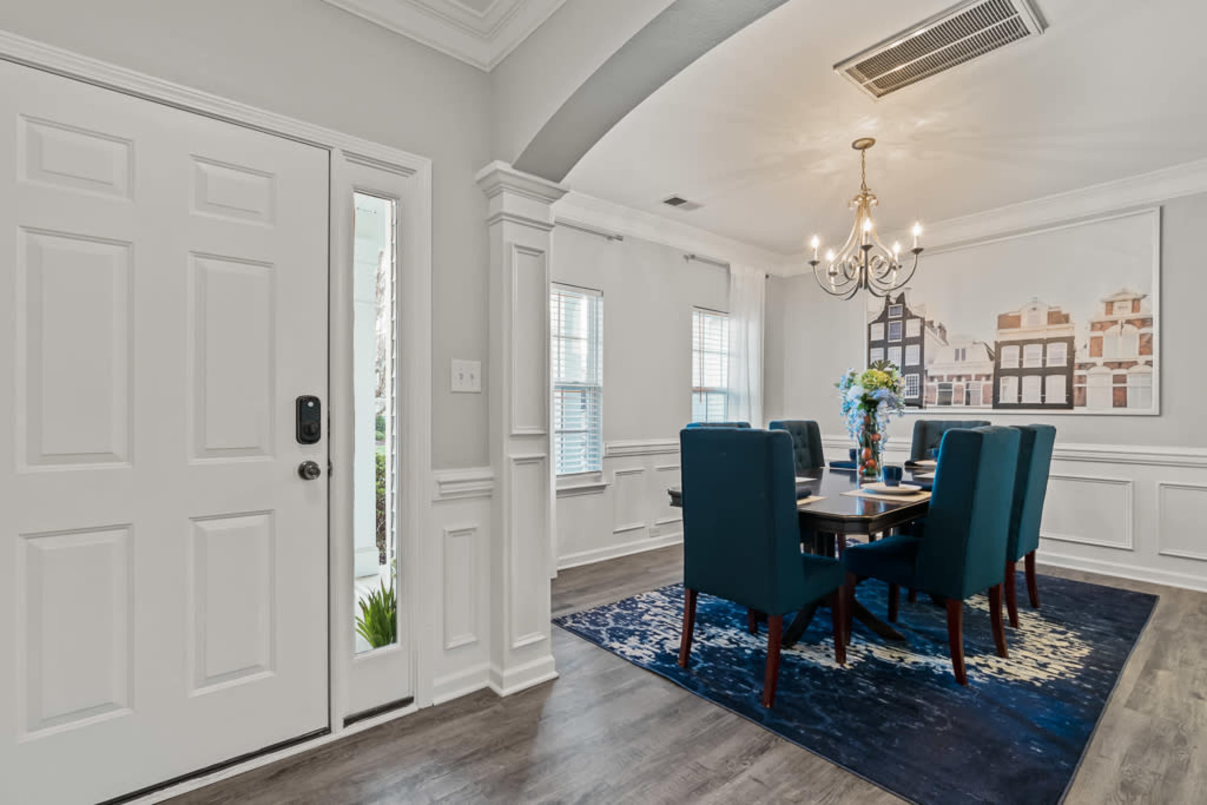 A dining area features a rectangular table surrounded by six teal chairs, with a large artwork on the wall and a chandelier overhead.