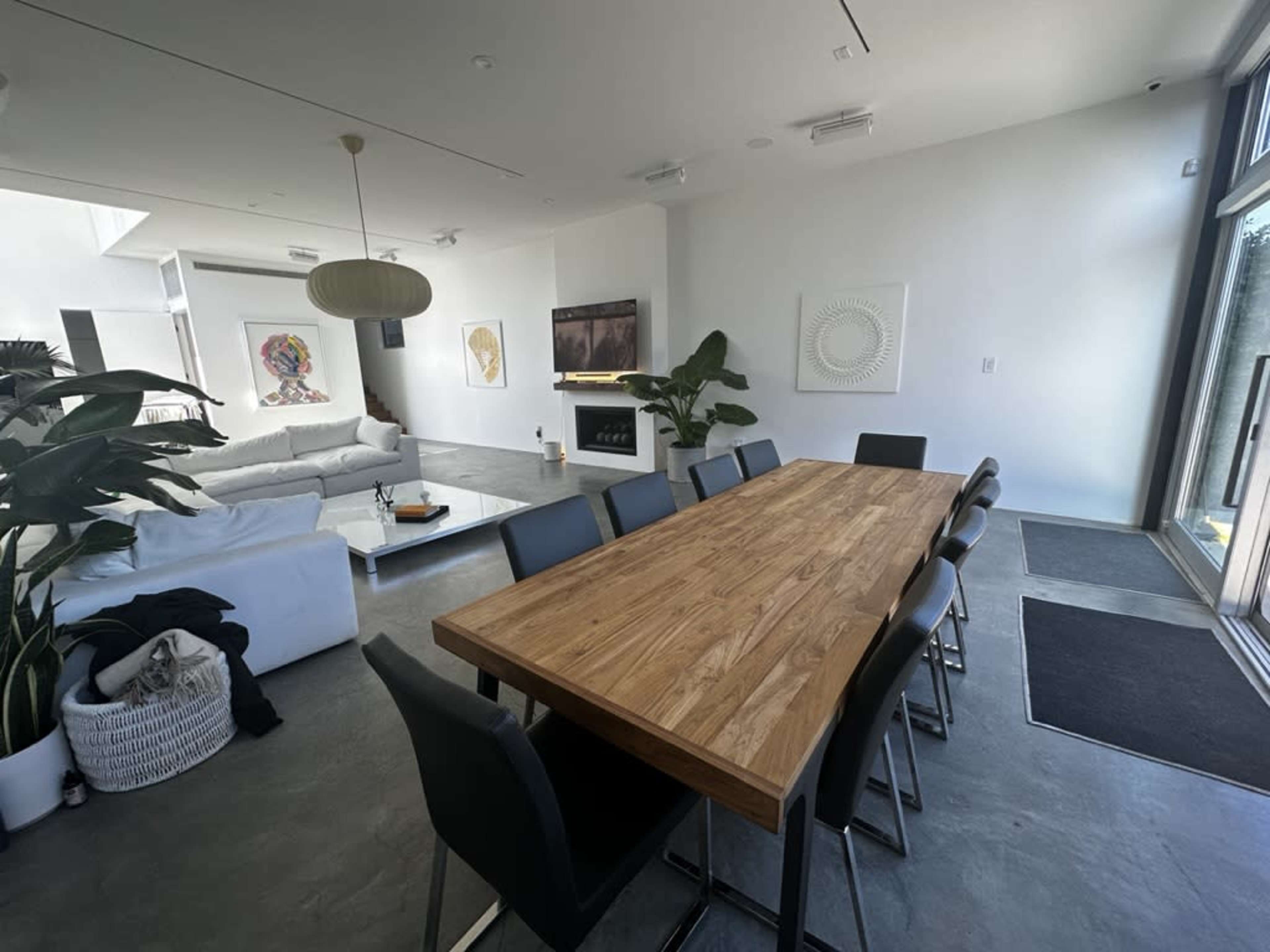 A spacious dining area features a long wooden table surrounded by black chairs, overlooking a modern living space with a white couch and large windows.