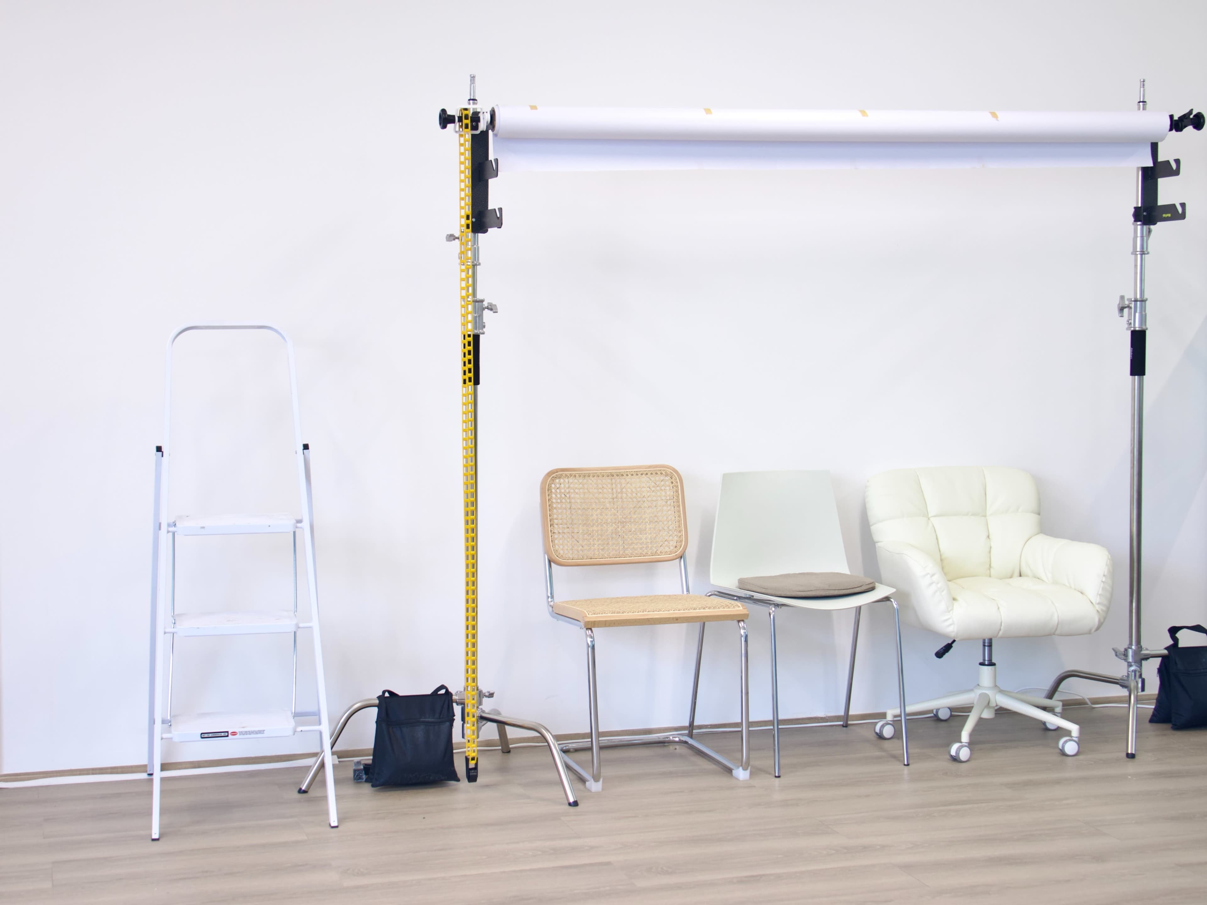 The image shows a simple studio setup featuring a ladder, a variety of chairs, and a backdrop support against a plain wall.