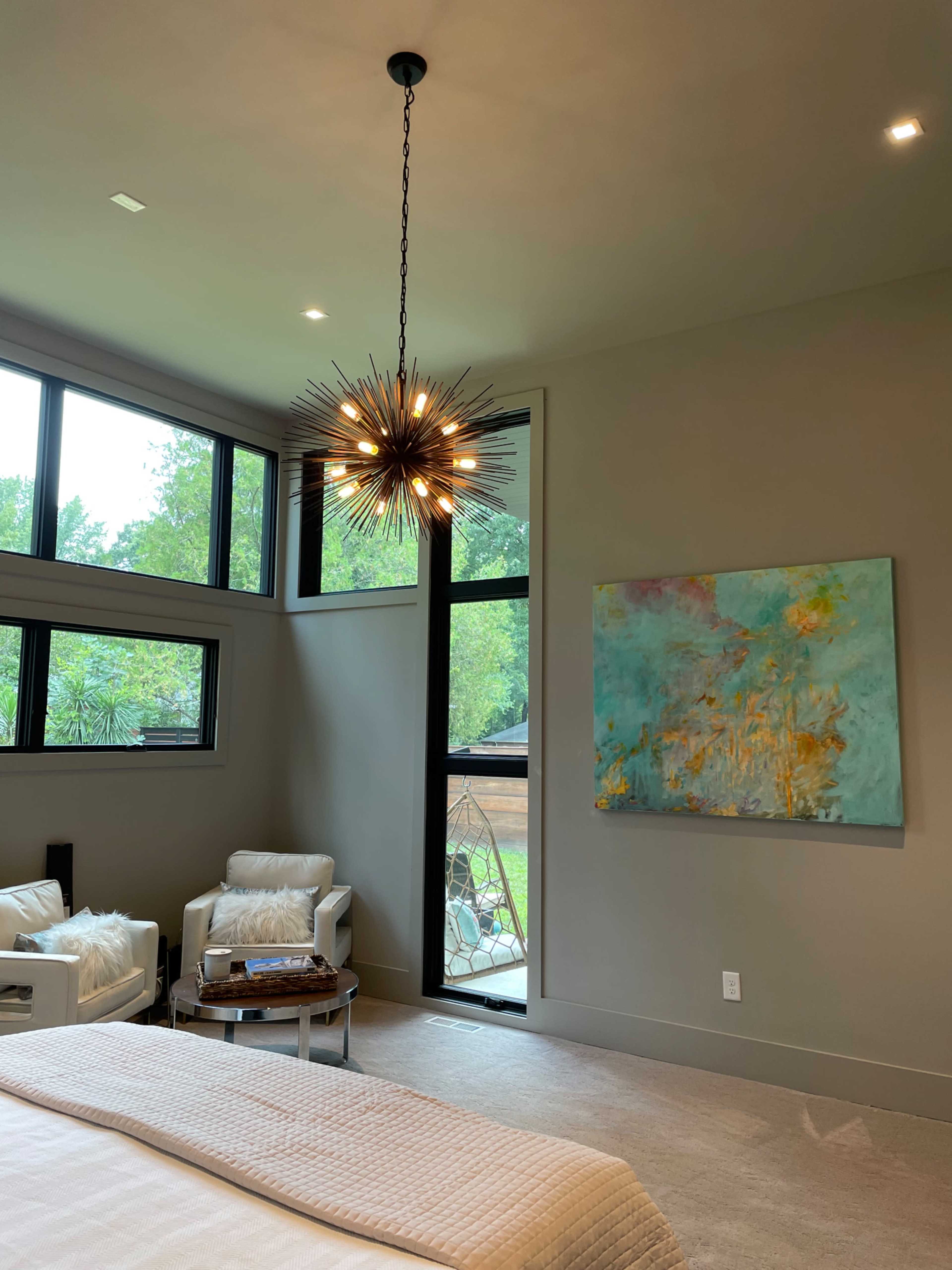 The image shows a modern bedroom featuring a starburst chandelier, a pair of armchairs, and a large abstract painting on the wall.