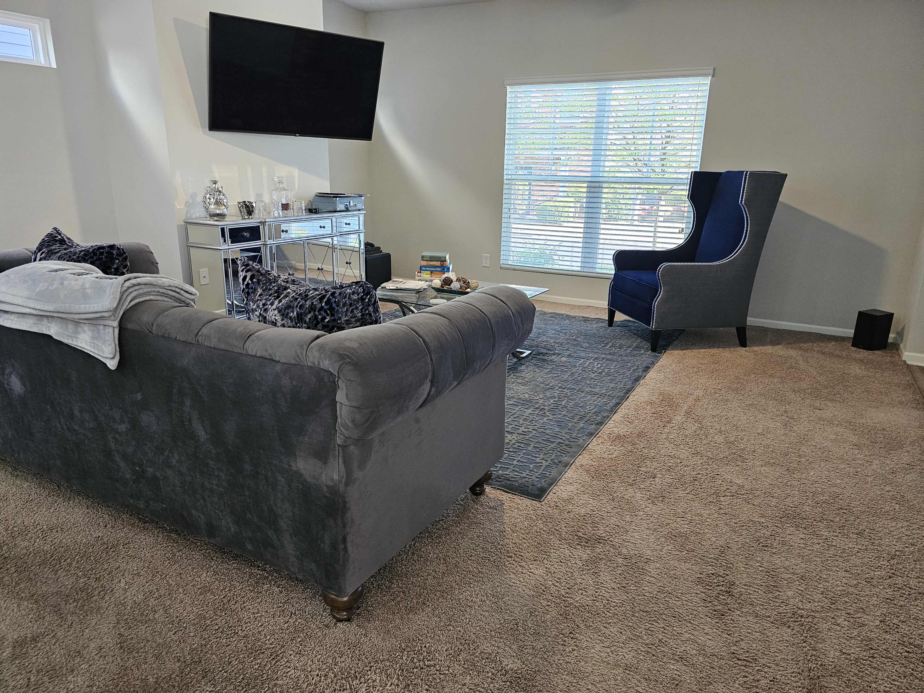 A cozy living room with a gray sofa, an armchair in the corner, a TV mounted on the wall, and a window with blinds allowing natural light to enter.