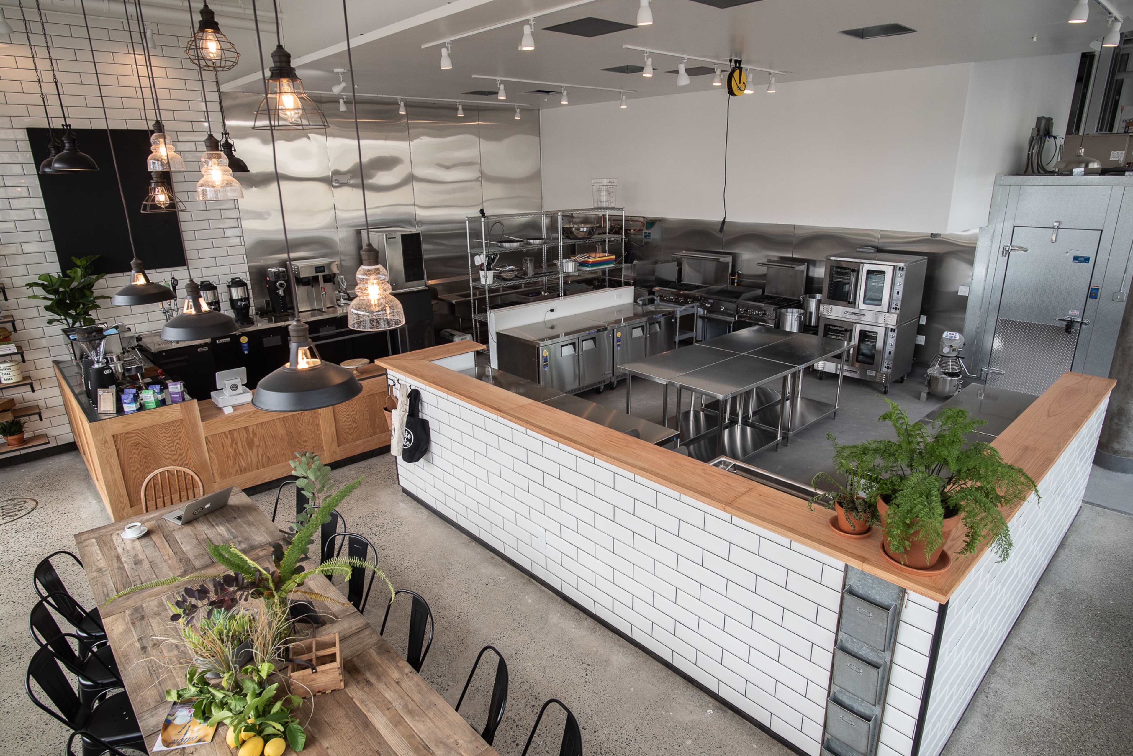 The image shows a modern industrial kitchen with stainless steel appliances and a large wooden dining table surrounded by chairs.