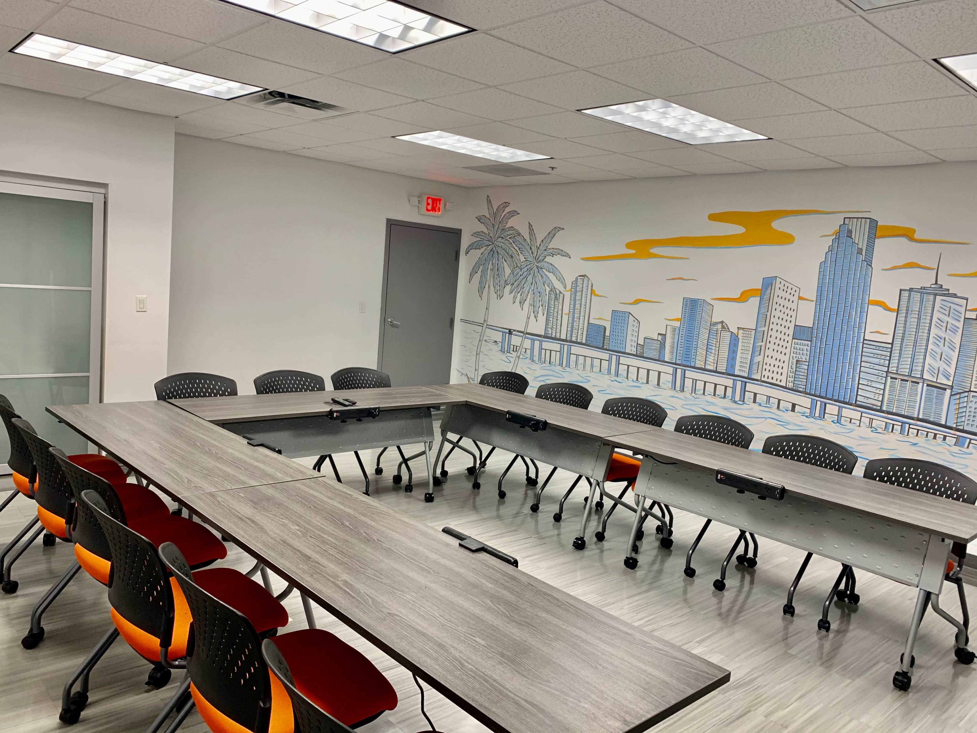 A conference room features a large, U-shaped table surrounded by colorful chairs, with a cityscape mural on one wall.