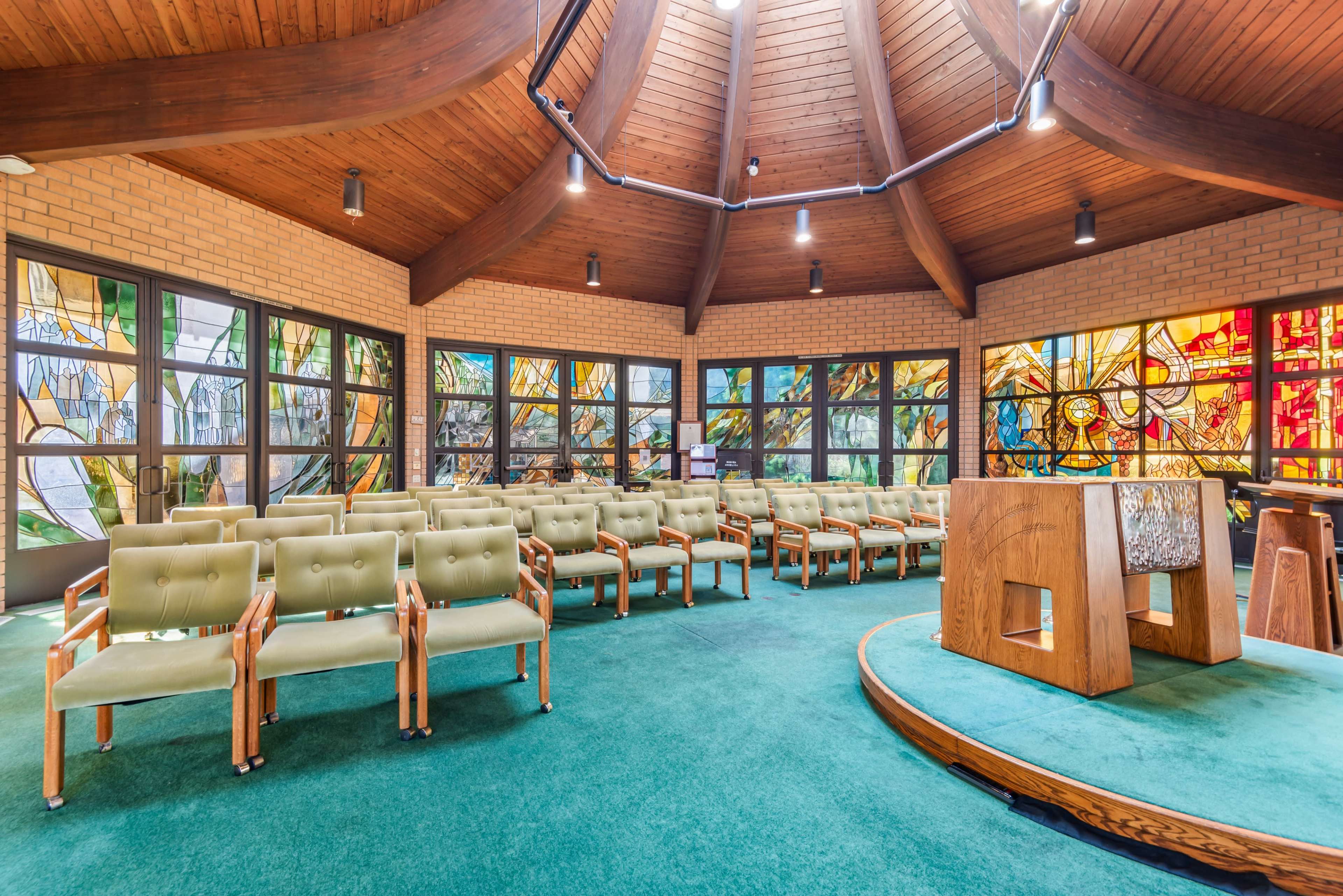 The image shows a spacious indoor room featuring rows of green upholstered chairs, a wooden altar, and large stained glass windows that let in natural light.