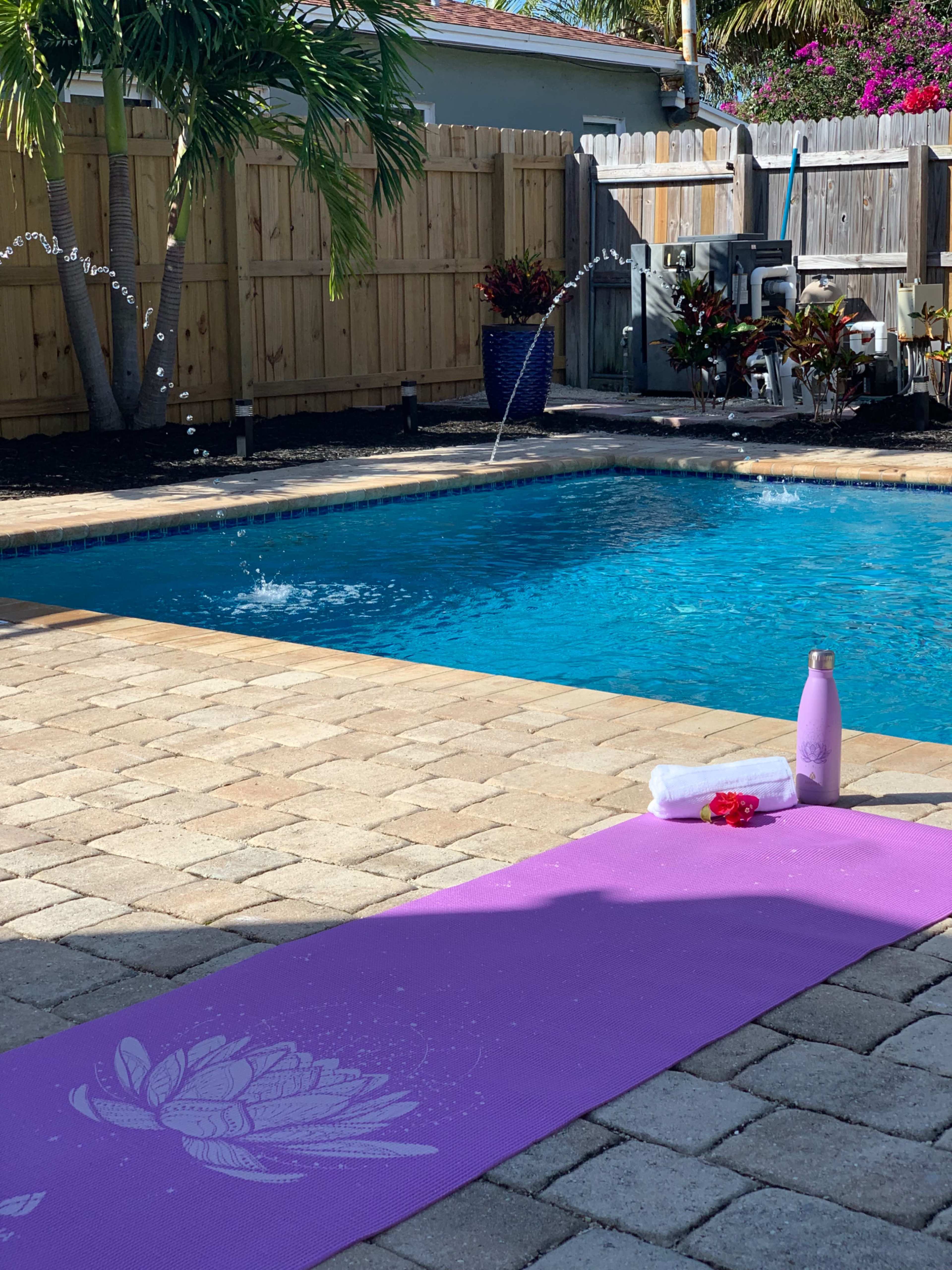 A yoga mat is placed on a stone patio beside a swimming pool, with a water bottle and a towel nearby.