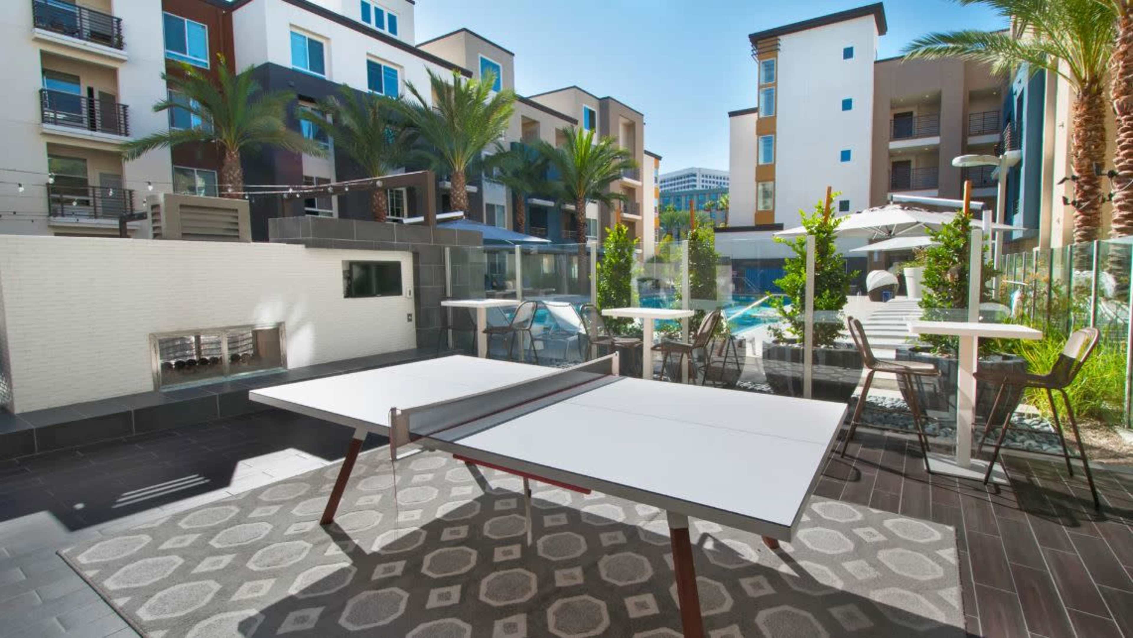 A modern outdoor area with a ping pong table, surrounded by palm trees, glass barriers, and seating areas near a pool.