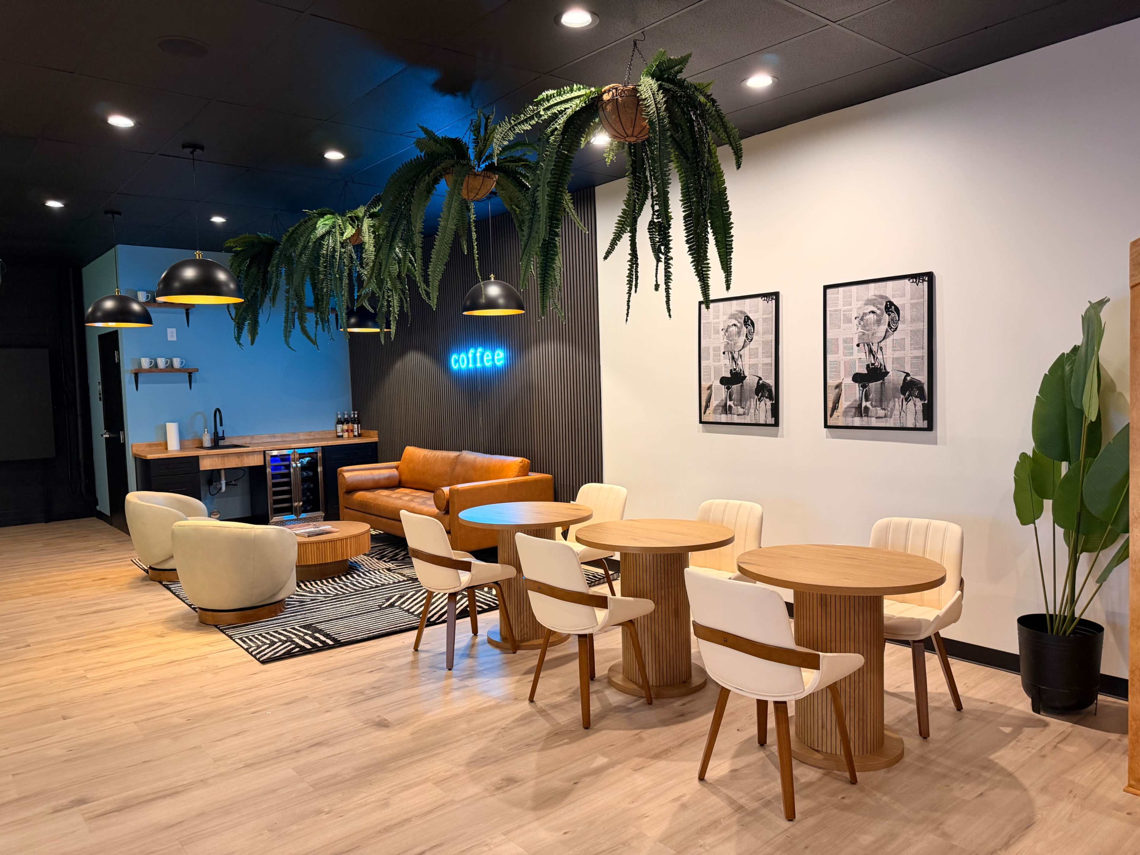 The image shows a modern cafe interior featuring a seating area with a brown leather couch, several wooden tables and chairs, and decorative plants hanging from the ceiling.