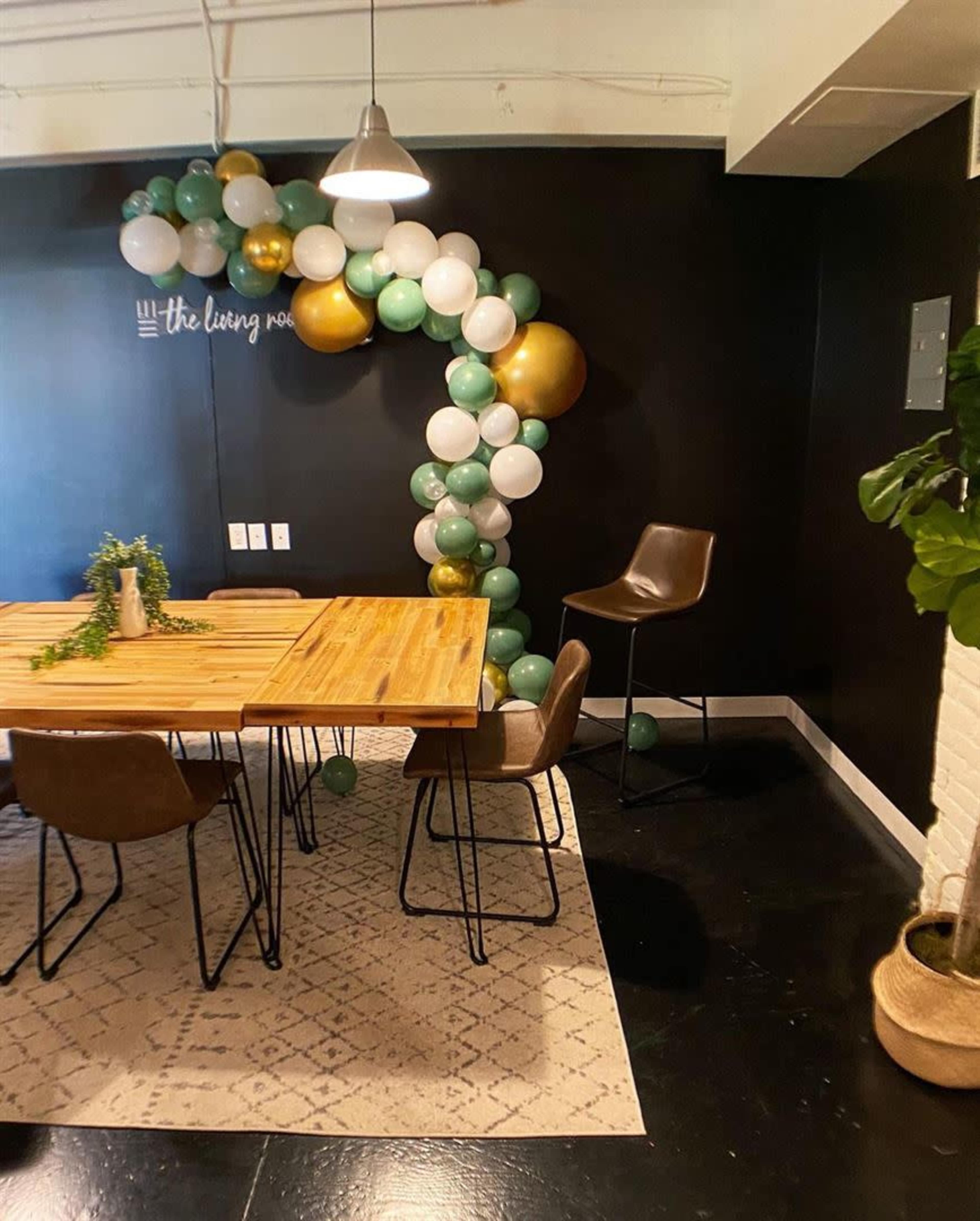 A dining area featuring a wooden table surrounded by chairs, with a decorative arch made of green, white, and gold balloons against a black wall.