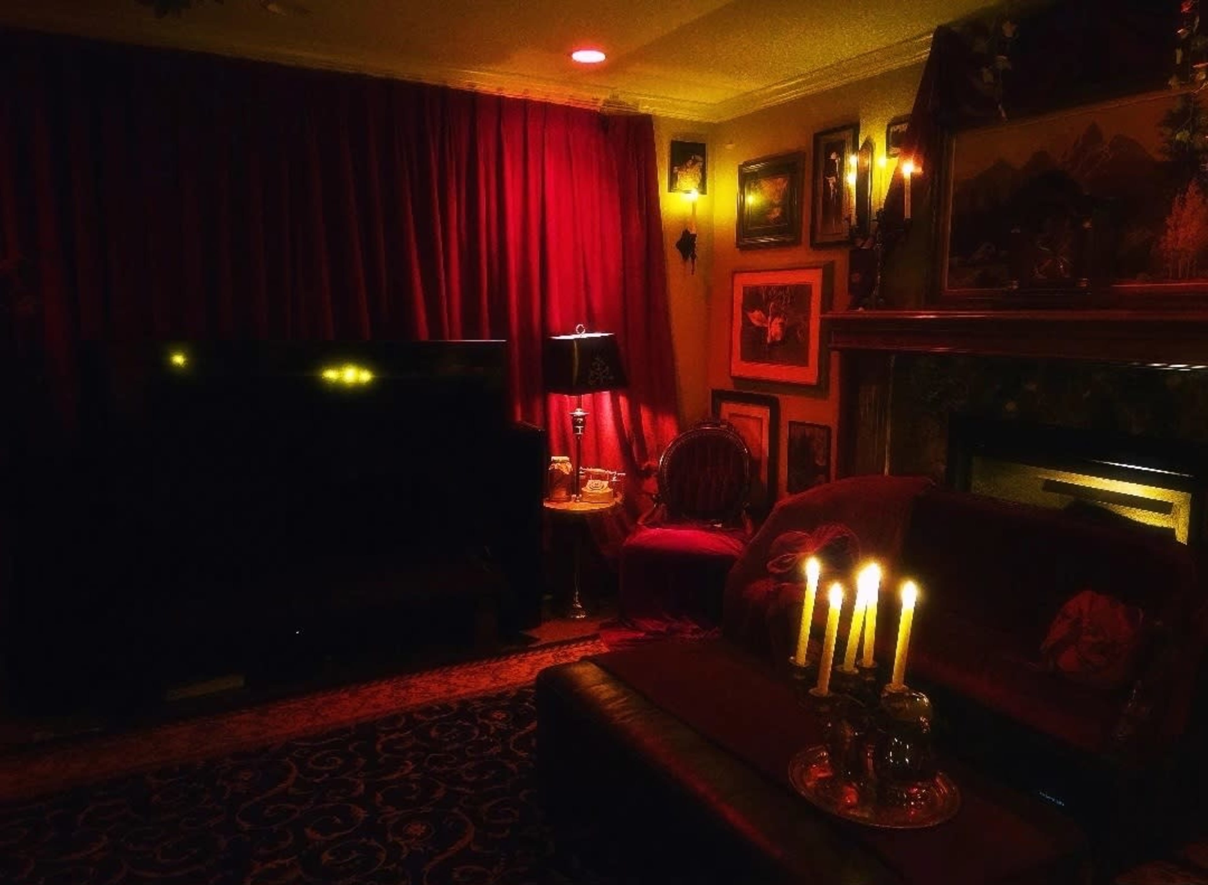 A dimly lit living room features red curtains, a television, candlelit decor, and framed artworks on the walls.