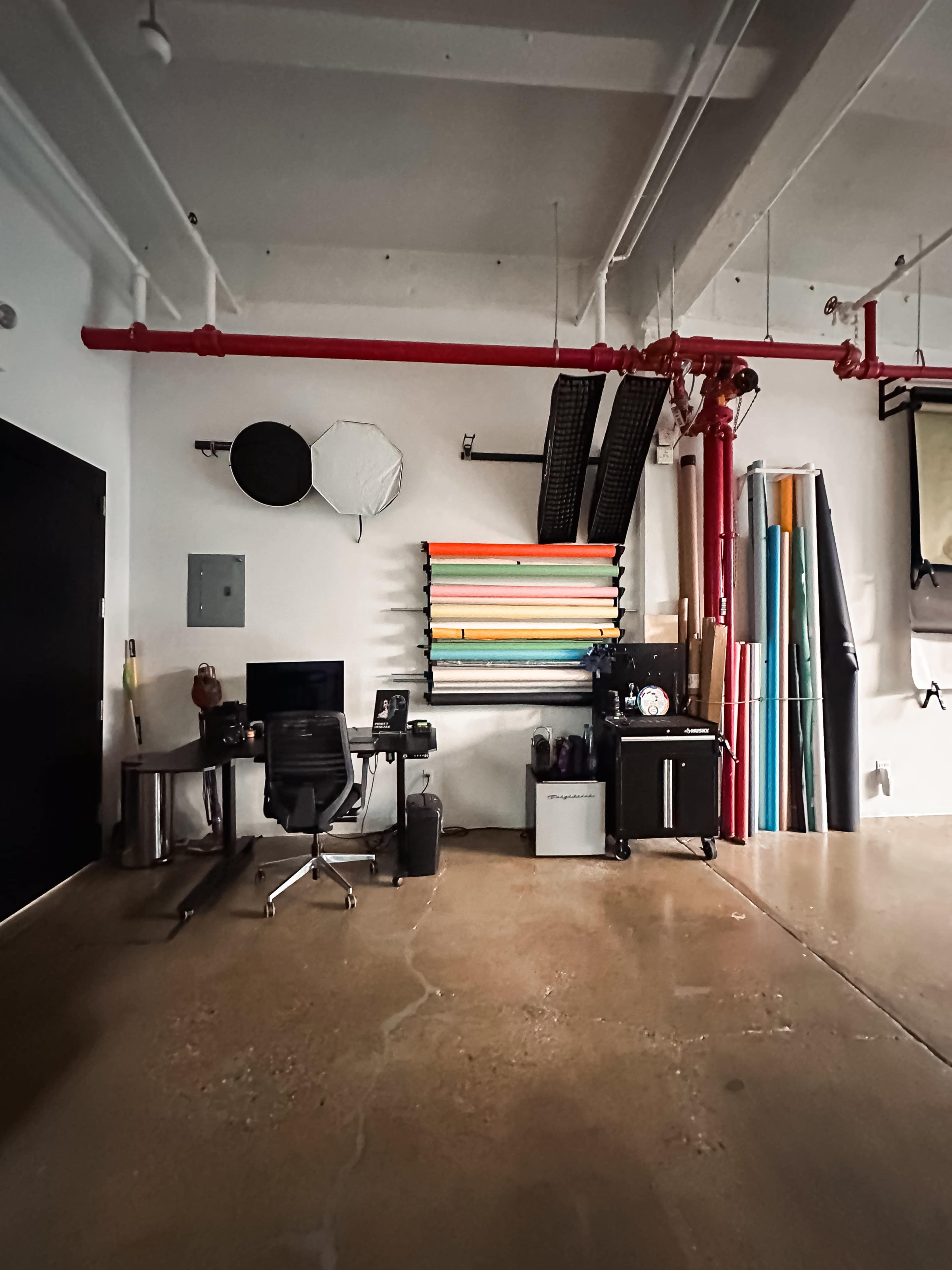 The scene shows a workspace featuring a desk with a chair, a variety of colorful backdrops stored vertically on a rack, and various studio equipment along a wall in a spacious room.