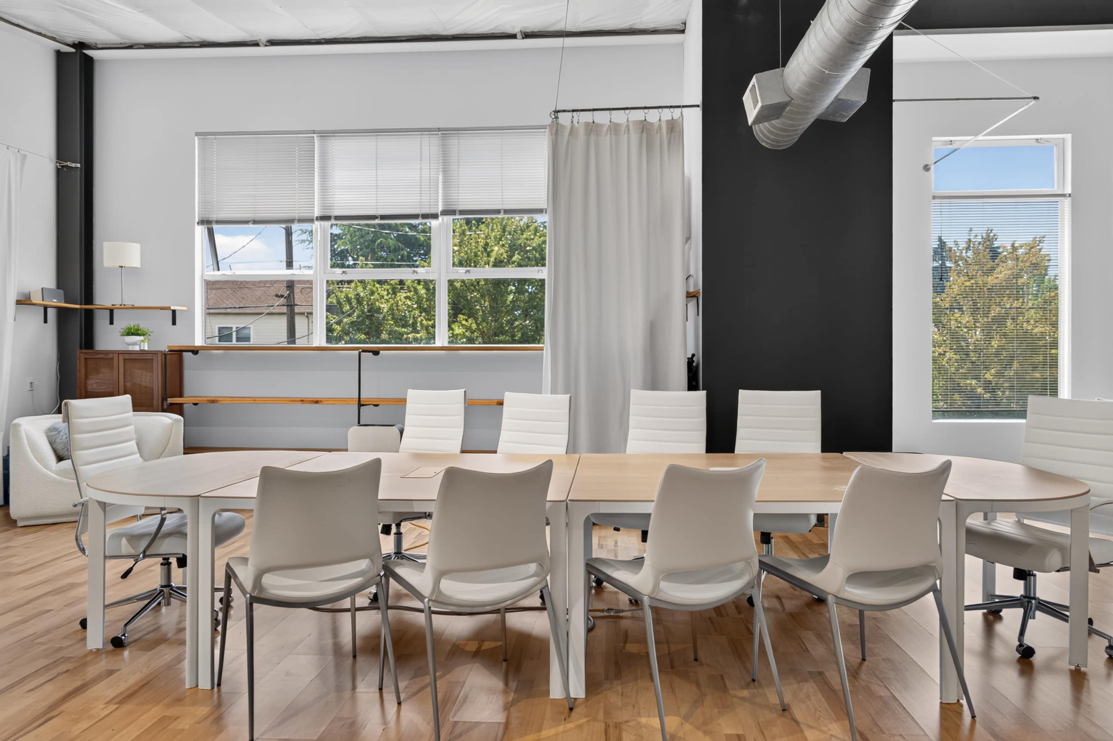 The image shows a bright conference room with a large wooden table surrounded by white chairs and large windows letting in natural light.