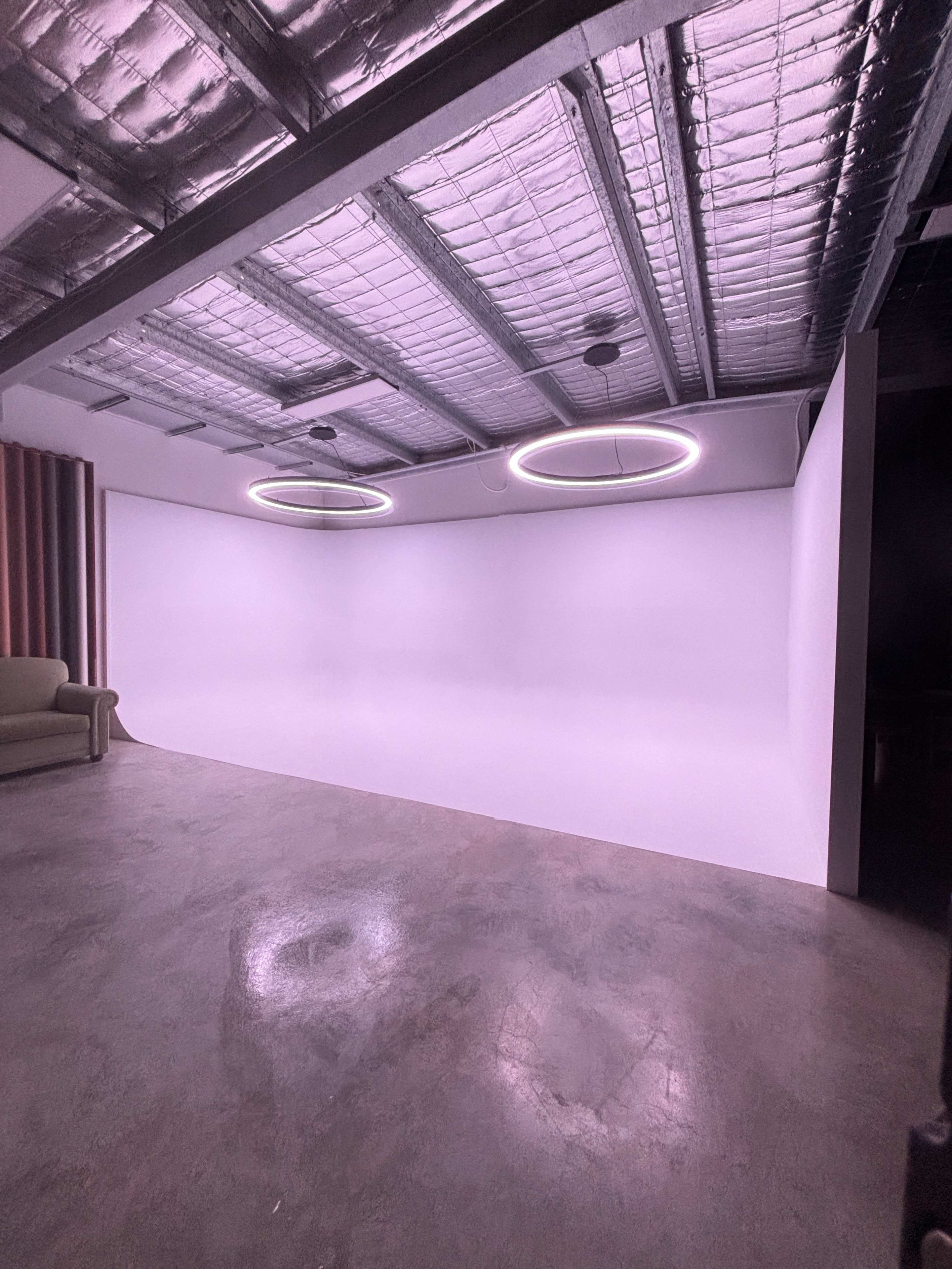 The image shows an empty room with a smooth white backdrop, two circular light fixtures on the ceiling, and a sofa against one wall.