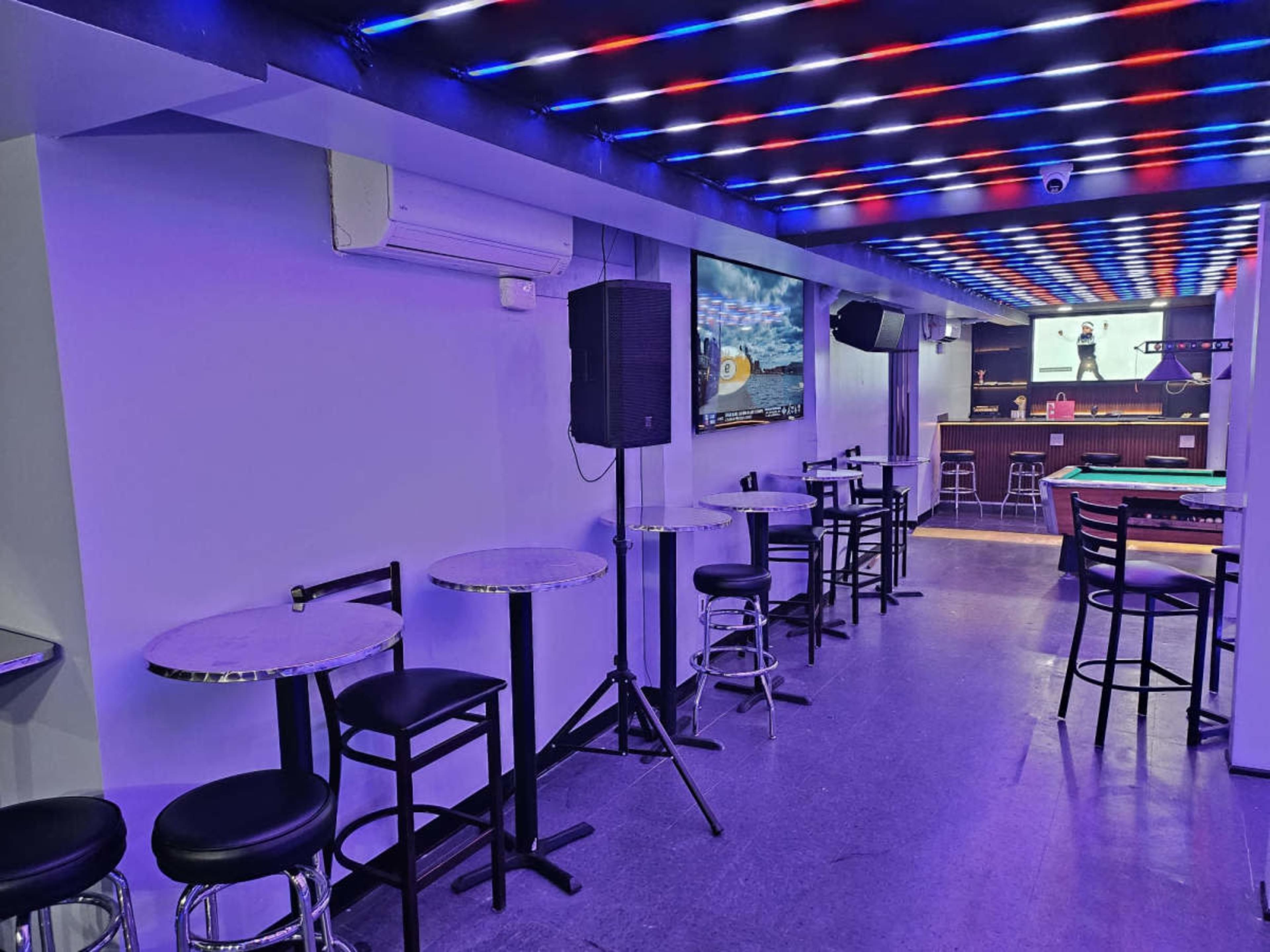 The image shows a modern lounge area with bar stools and small tables under colorful LED lighting and screens on the walls.