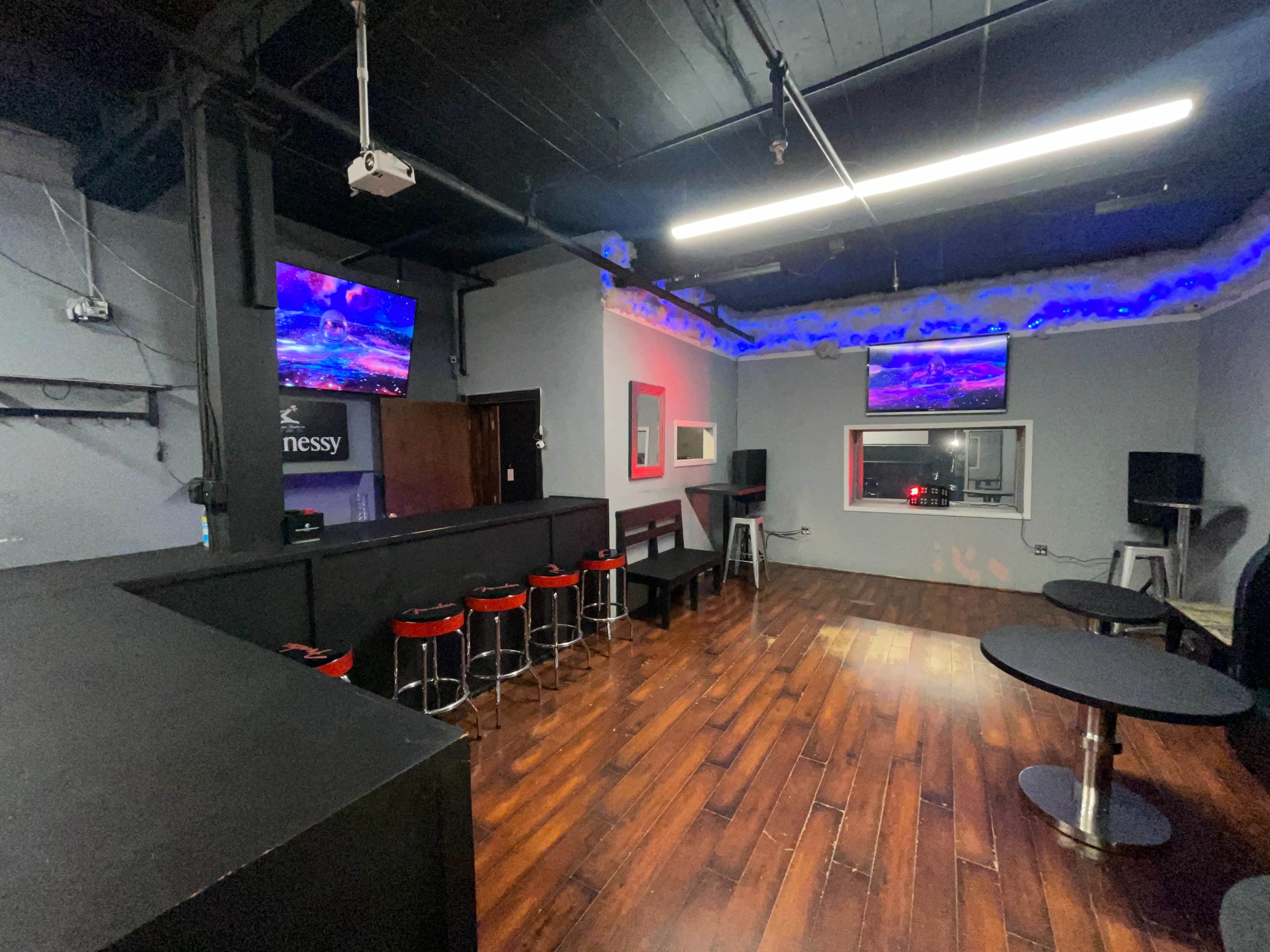 A dimly lit bar or lounge area featuring dark wooden floors, high-top tables, and two wall-mounted televisions displaying colorful visuals.