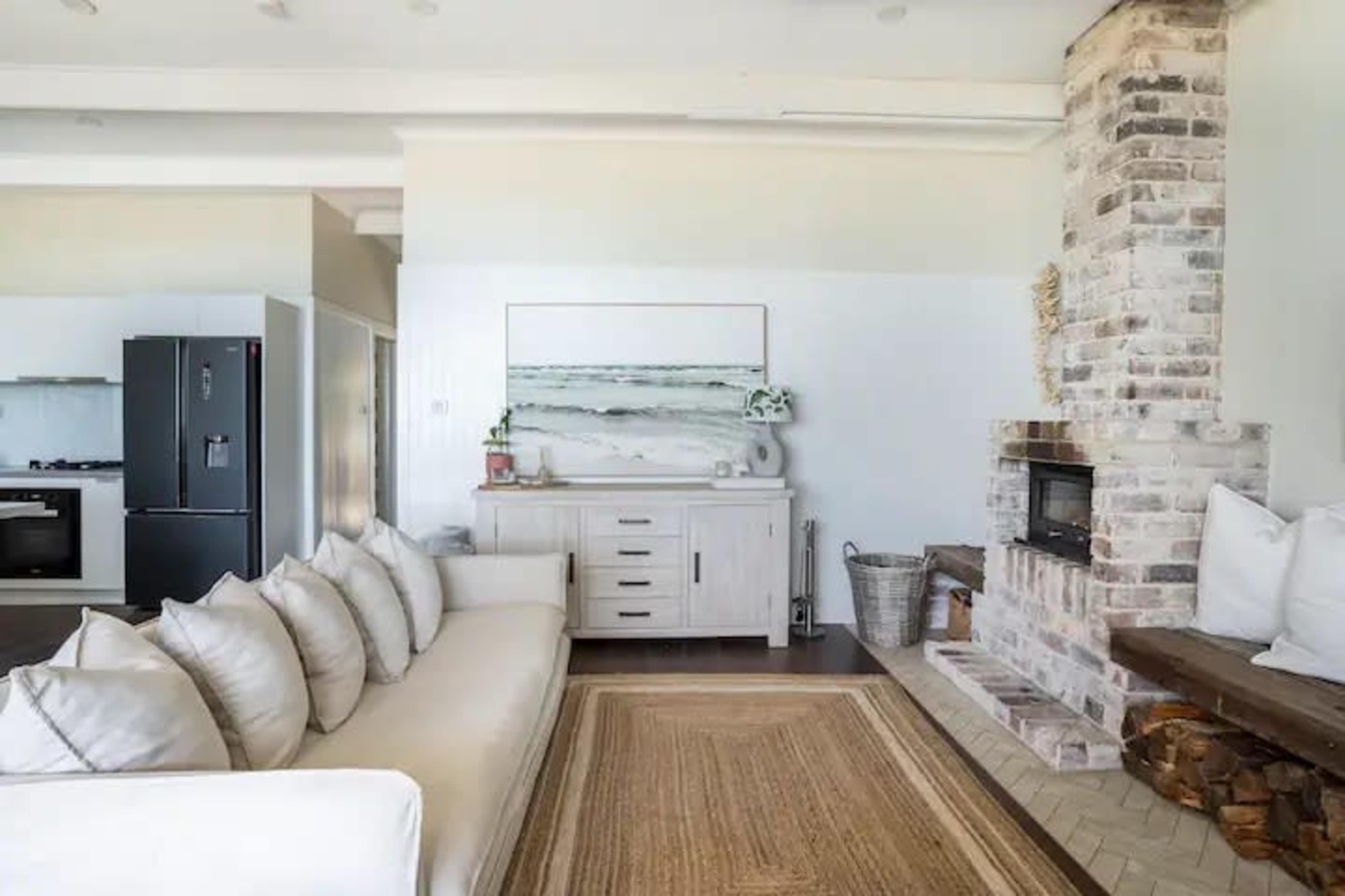 A living room featuring a white sofa with multiple cushions, a wooden sideboard, a brick fireplace, and a textured rug on the floor.