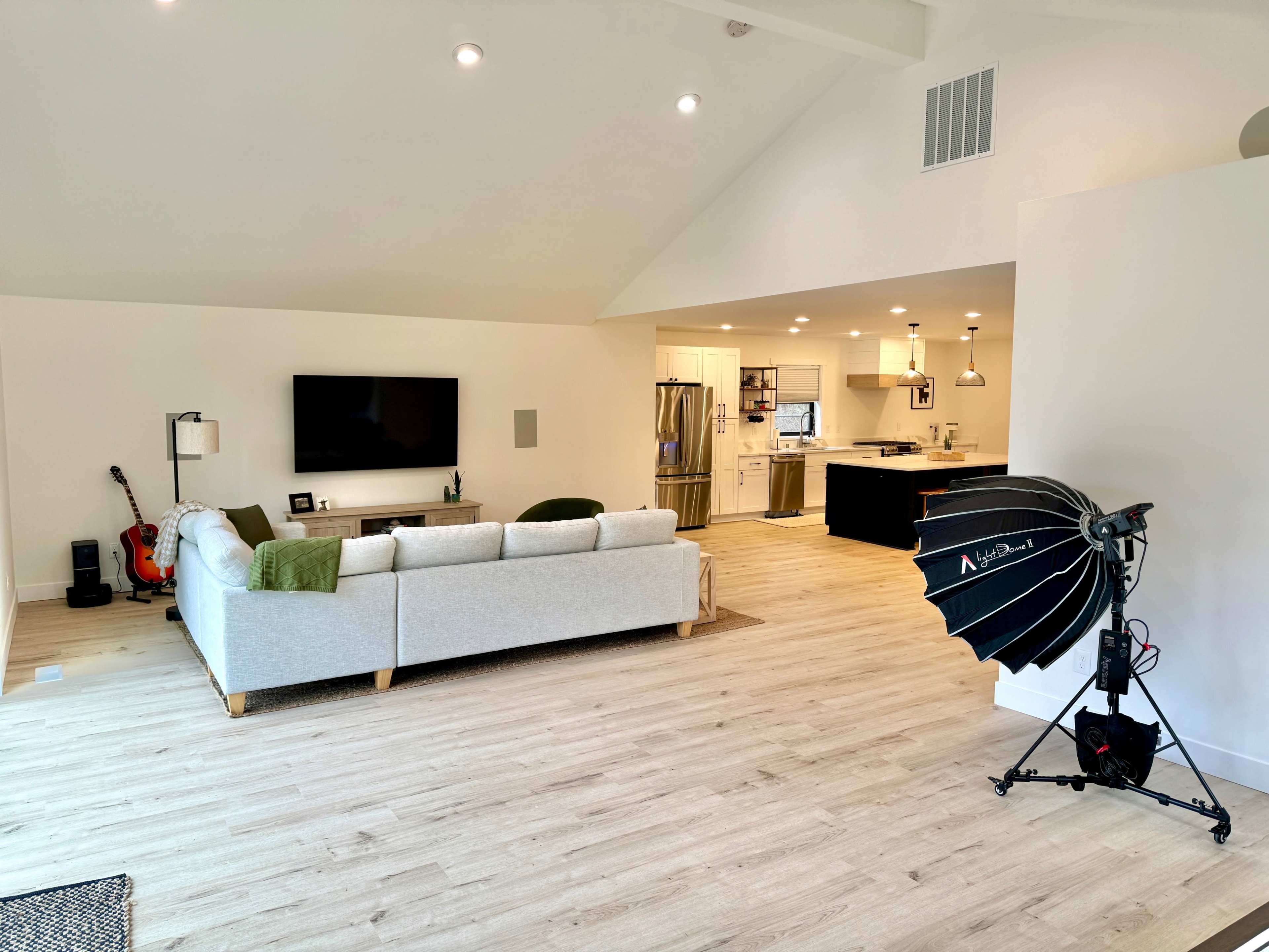 The image shows a spacious living room with a light-colored sofa, a television mounted on the wall, and an open kitchen visible in the background.