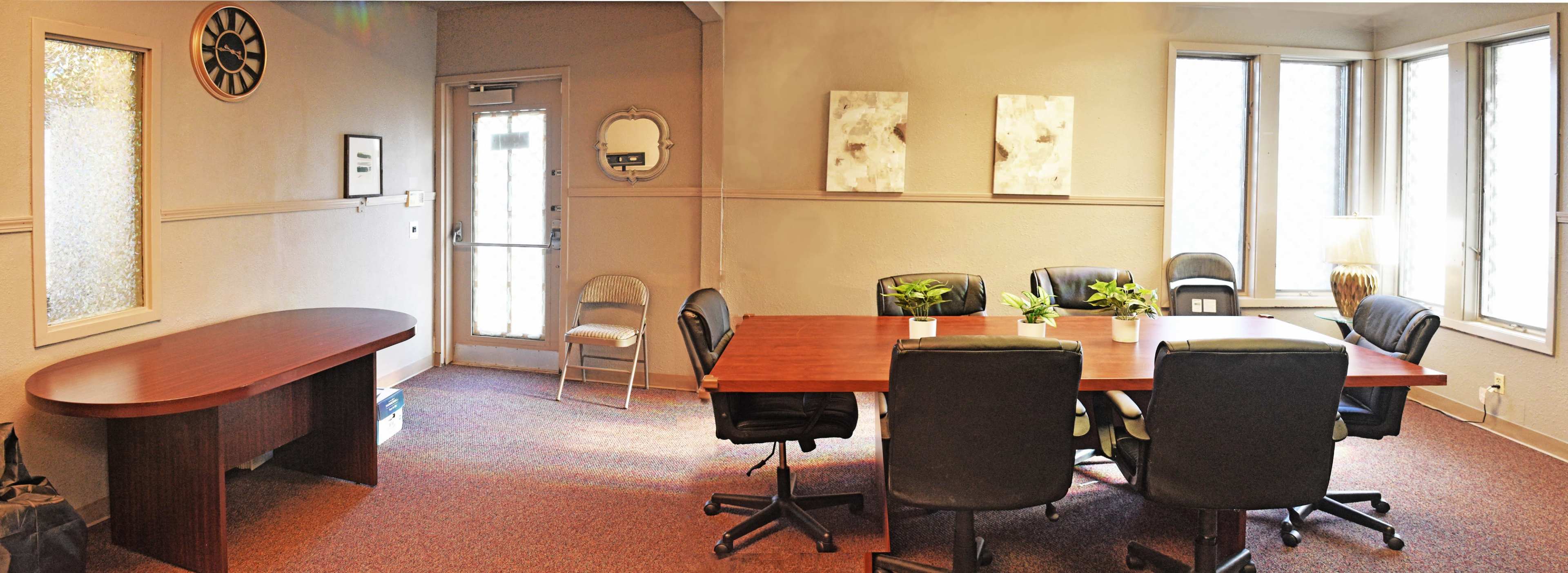 A conference room with a large wooden table surrounded by black office chairs, a waiting area with a folding chair, and large windows allowing natural light.