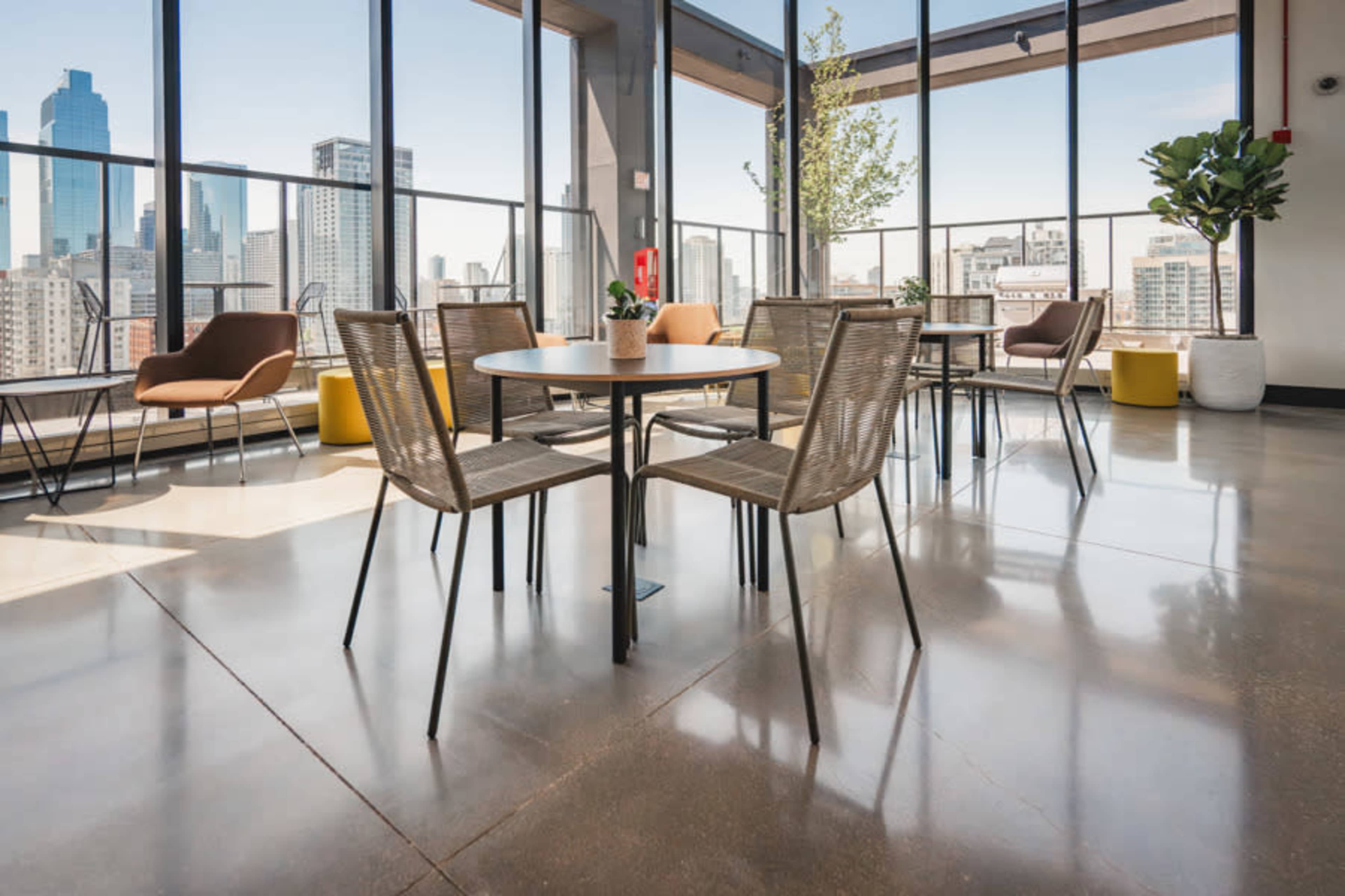 The image shows a modern, well-lit lounge area with tables and chairs arranged near large windows overlooking a city skyline.