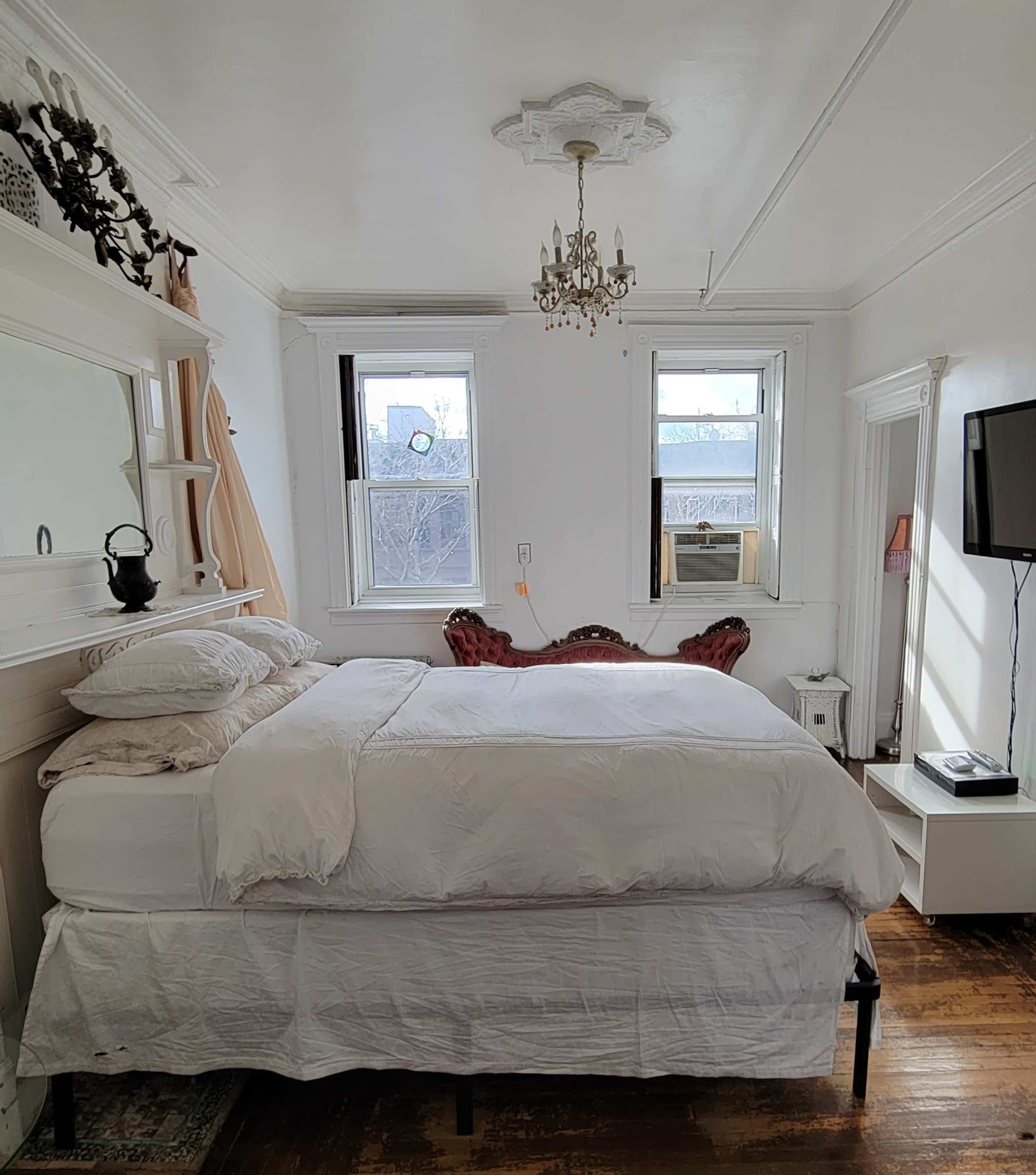 A bedroom features a bed with white bedding, a vintage chandelier, two windows, a television on a stand, and a plush seating area.