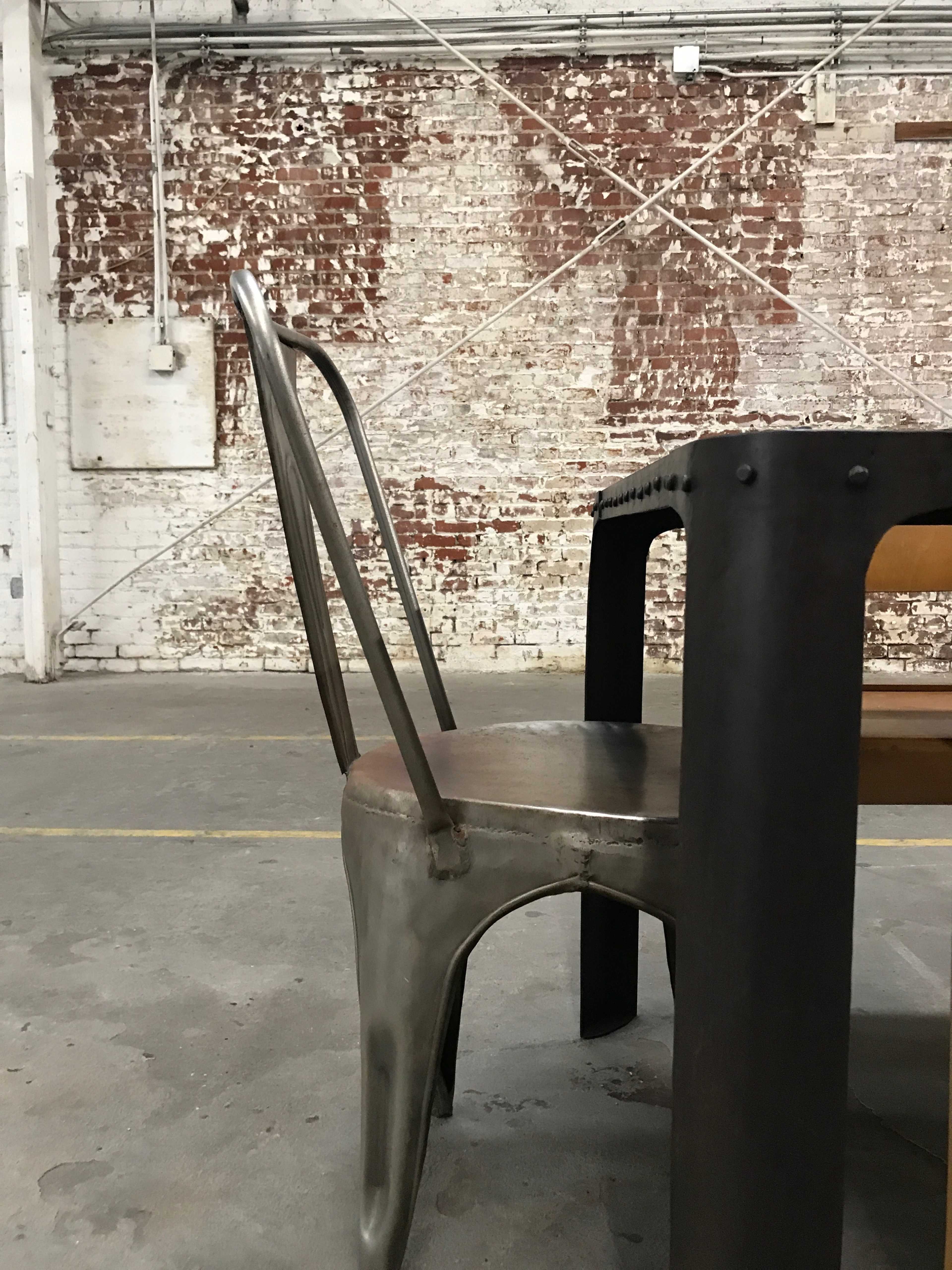 A metal chair stands next to a wooden table against a textured, brick wall in an industrial space.