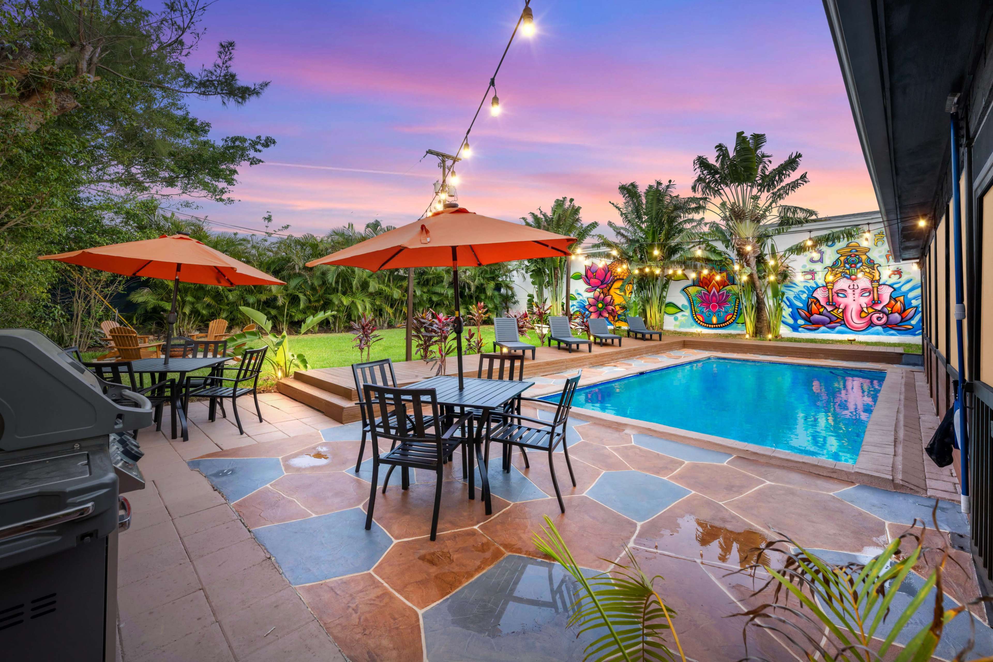 A backyard with a swimming pool, surrounded by patio furniture under orange umbrellas, and a vibrant mural on the wall.