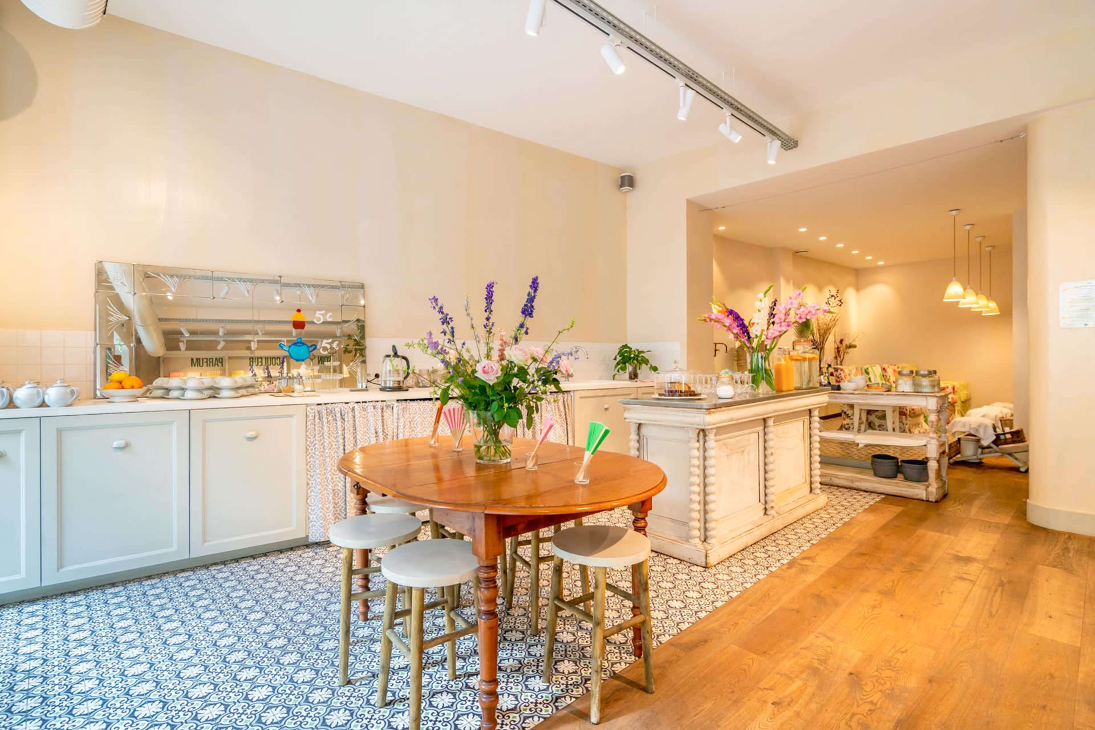 A bright kitchen and dining area features a round wooden table surrounded by chairs, with floral arrangements and a side counter displaying food and drinks.