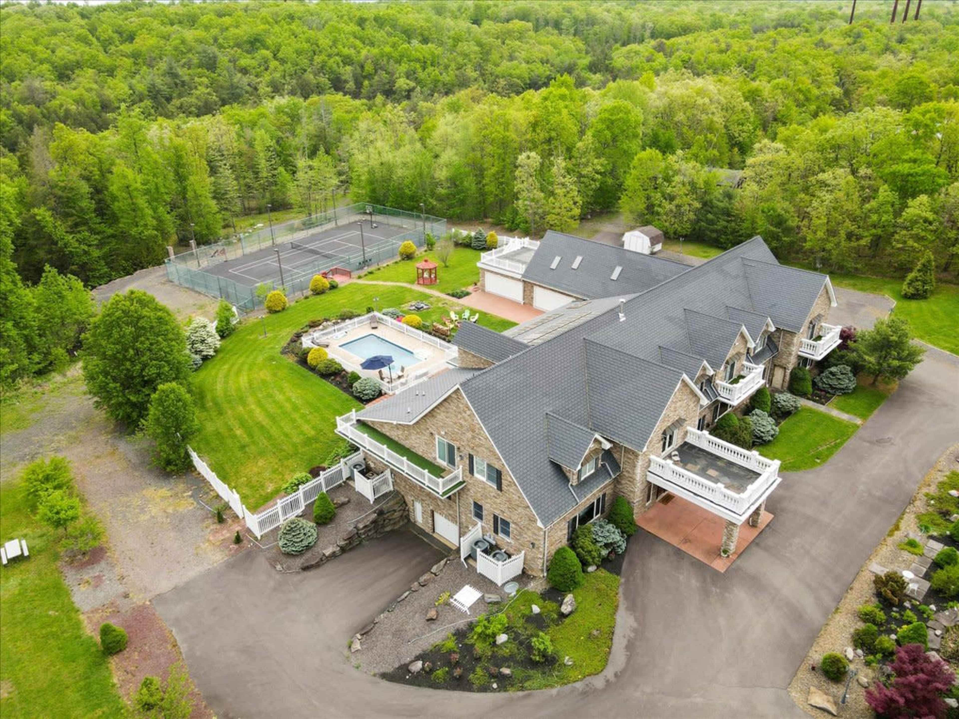 A large stone house with a swimming pool and tennis court surrounded by lush greenery.
