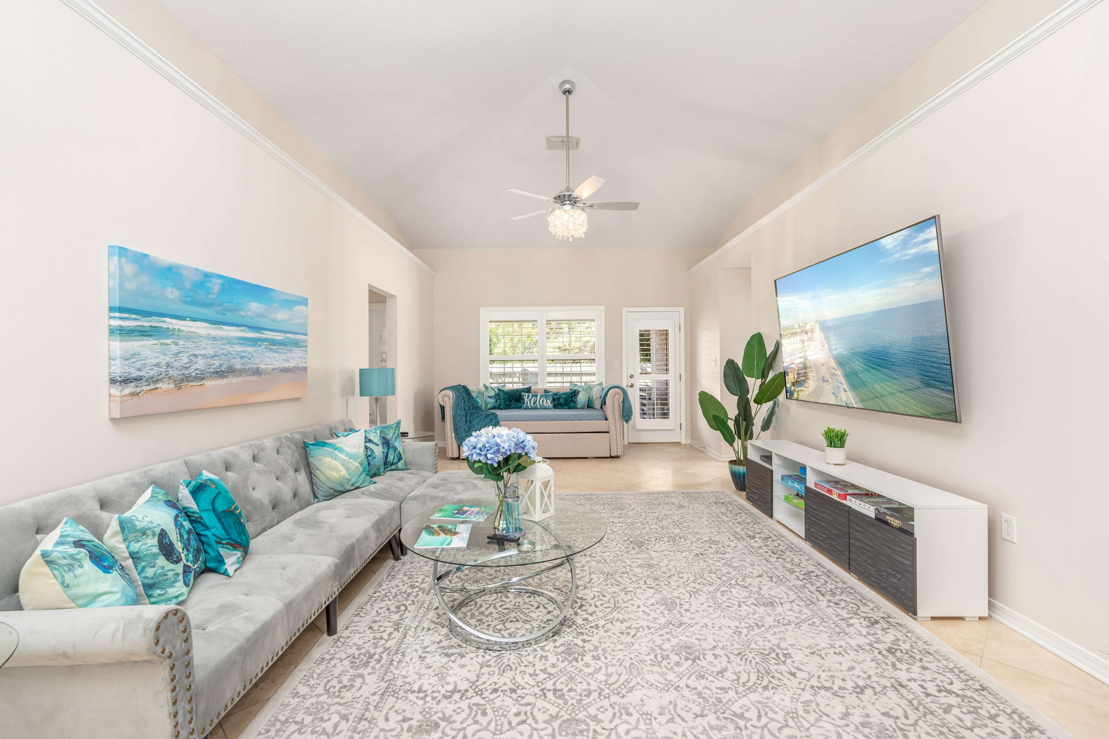 A living room features a gray sofa, a glass coffee table, a large flat-screen TV mounted on the wall, and decorative ocean-themed artwork.