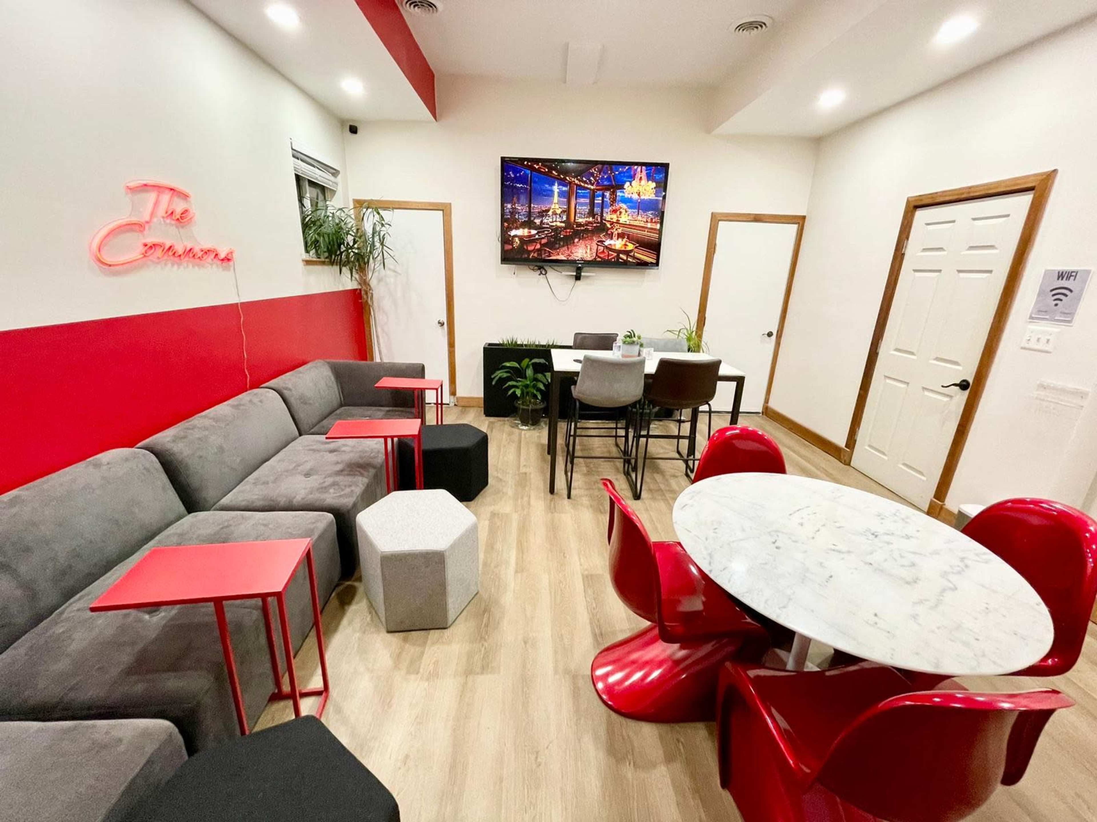 A modern room featuring a gray sectional sofa, a round marble table with red chairs, and a wall-mounted TV displaying a cityscape.