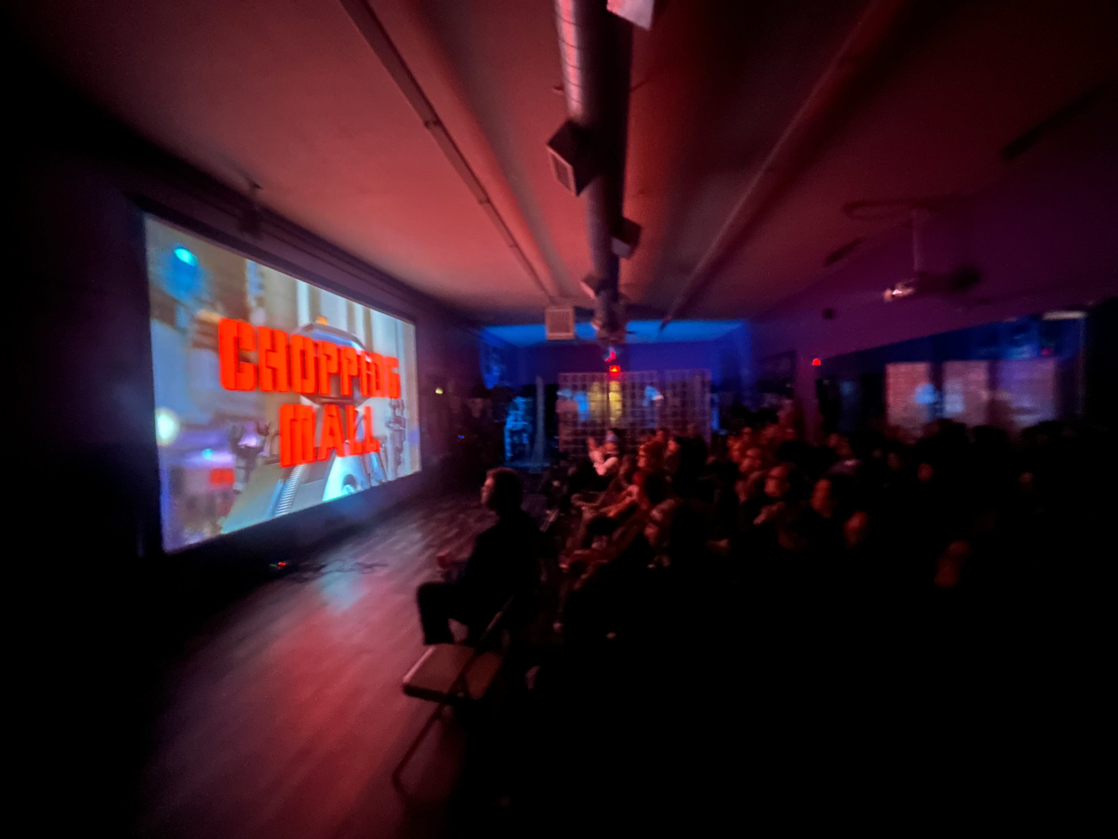 A crowd sits in a dimly lit room watching a large screen displaying the title "Chopping Mall."
