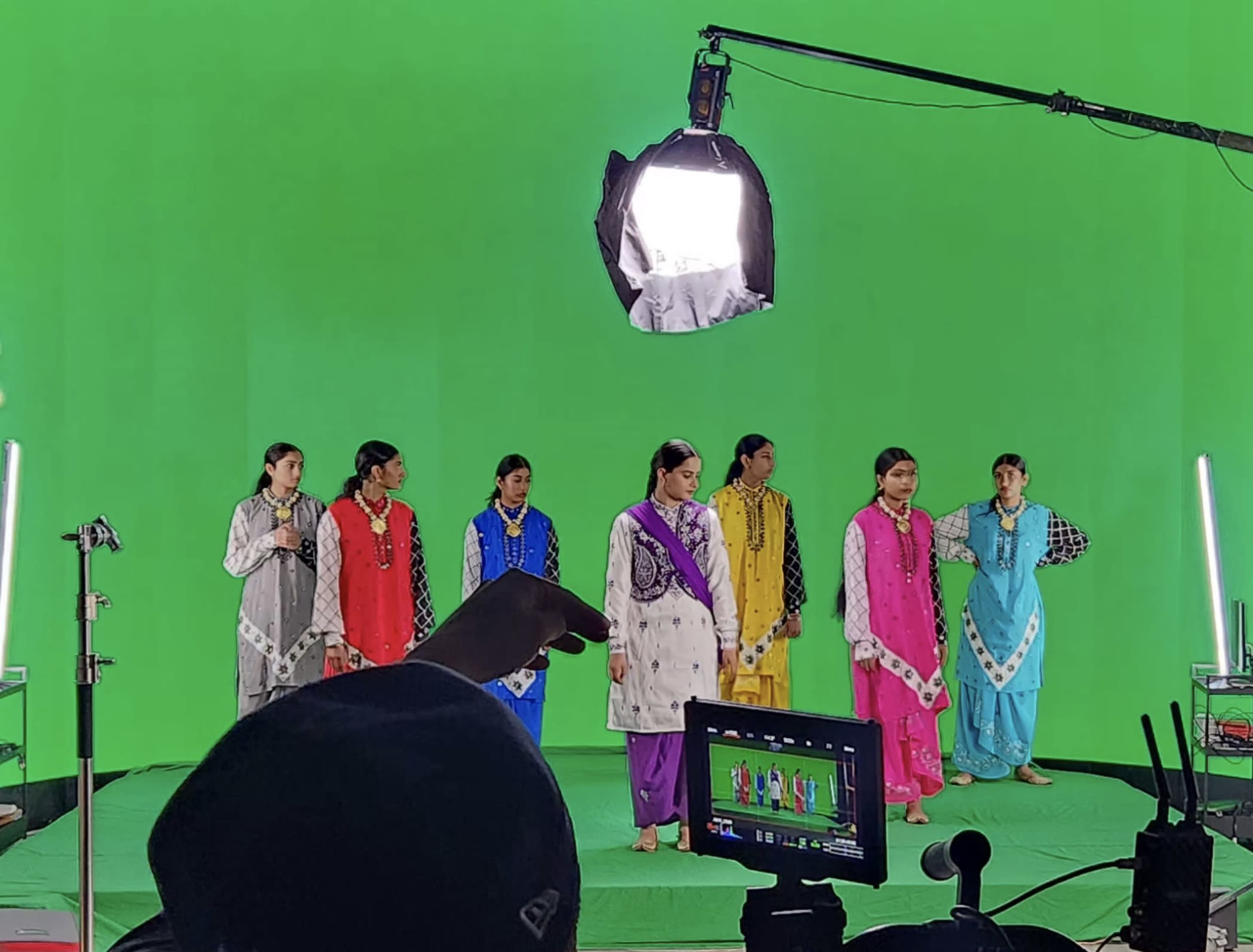 A group of six dancers, dressed in colorful traditional outfits, stands in front of a green screen on a film set.