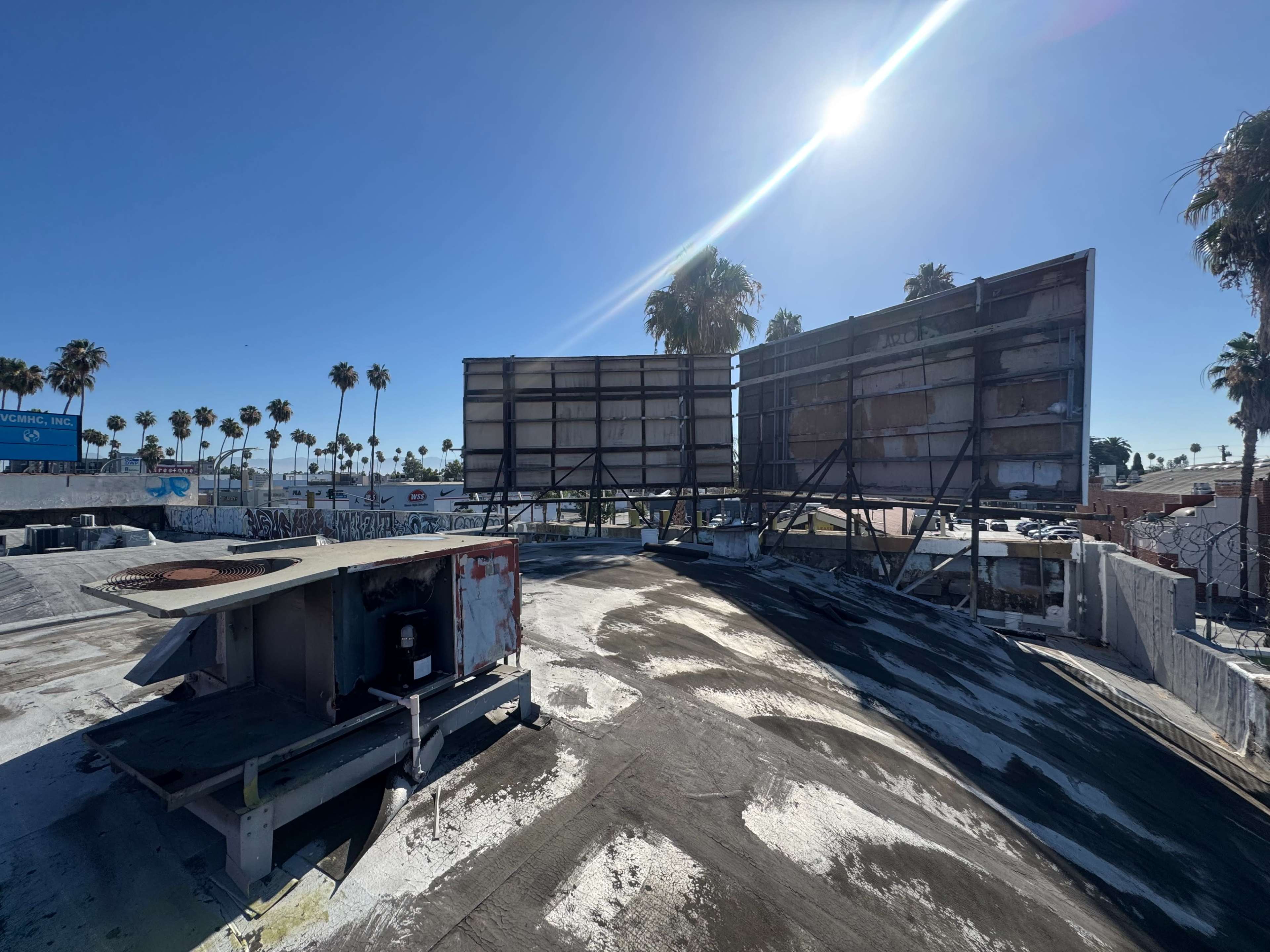 The image shows two large billboards on a rooftop surrounded by palm trees and a clear blue sky.