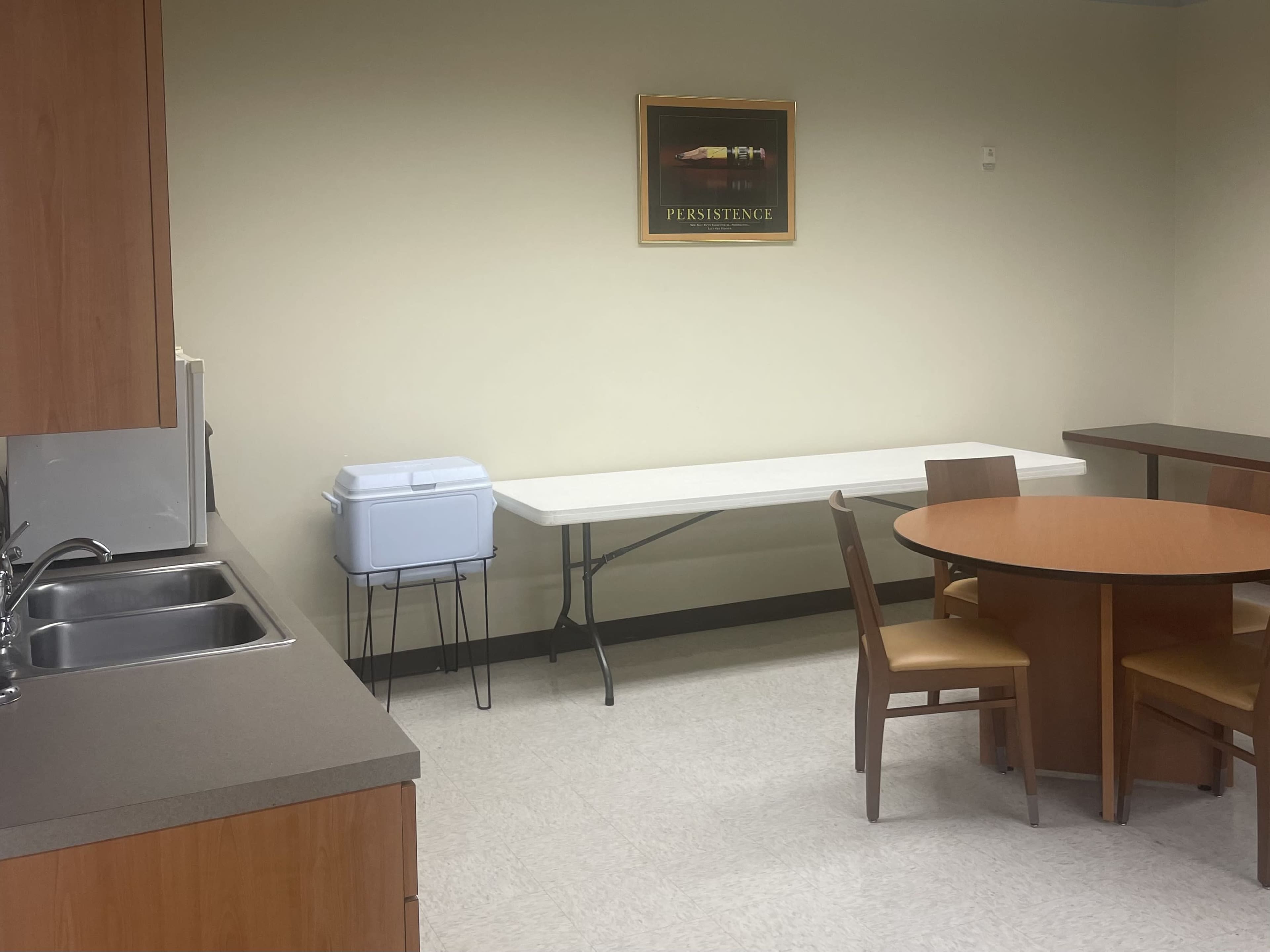 A small kitchen area with a sink and a table surrounded by chairs, along with a white folding table and a framed picture on the wall.