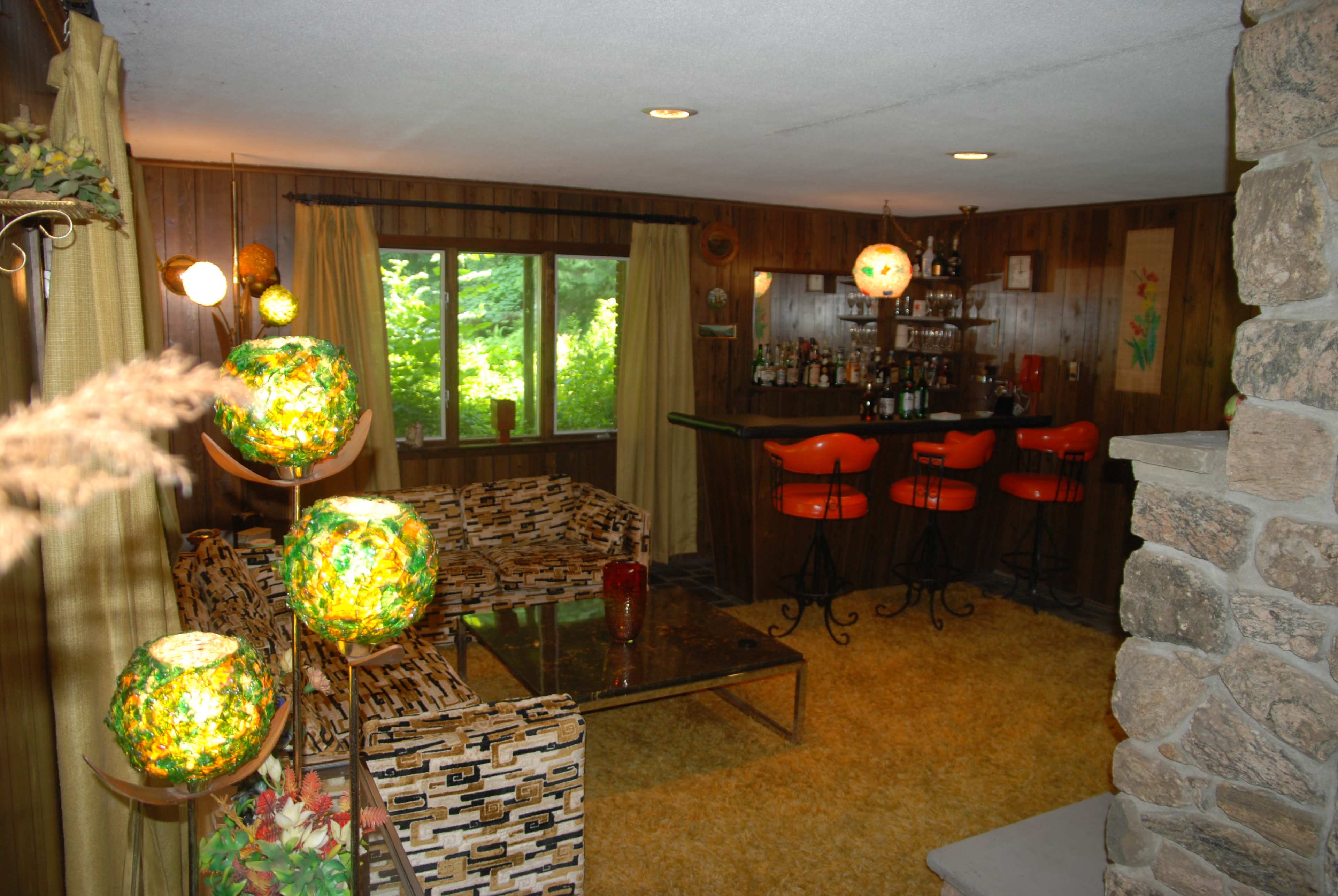 The image shows a retro-styled living room with wooden walls, a bar area, and orange stools, featuring a patterned couch and decorative lighting.