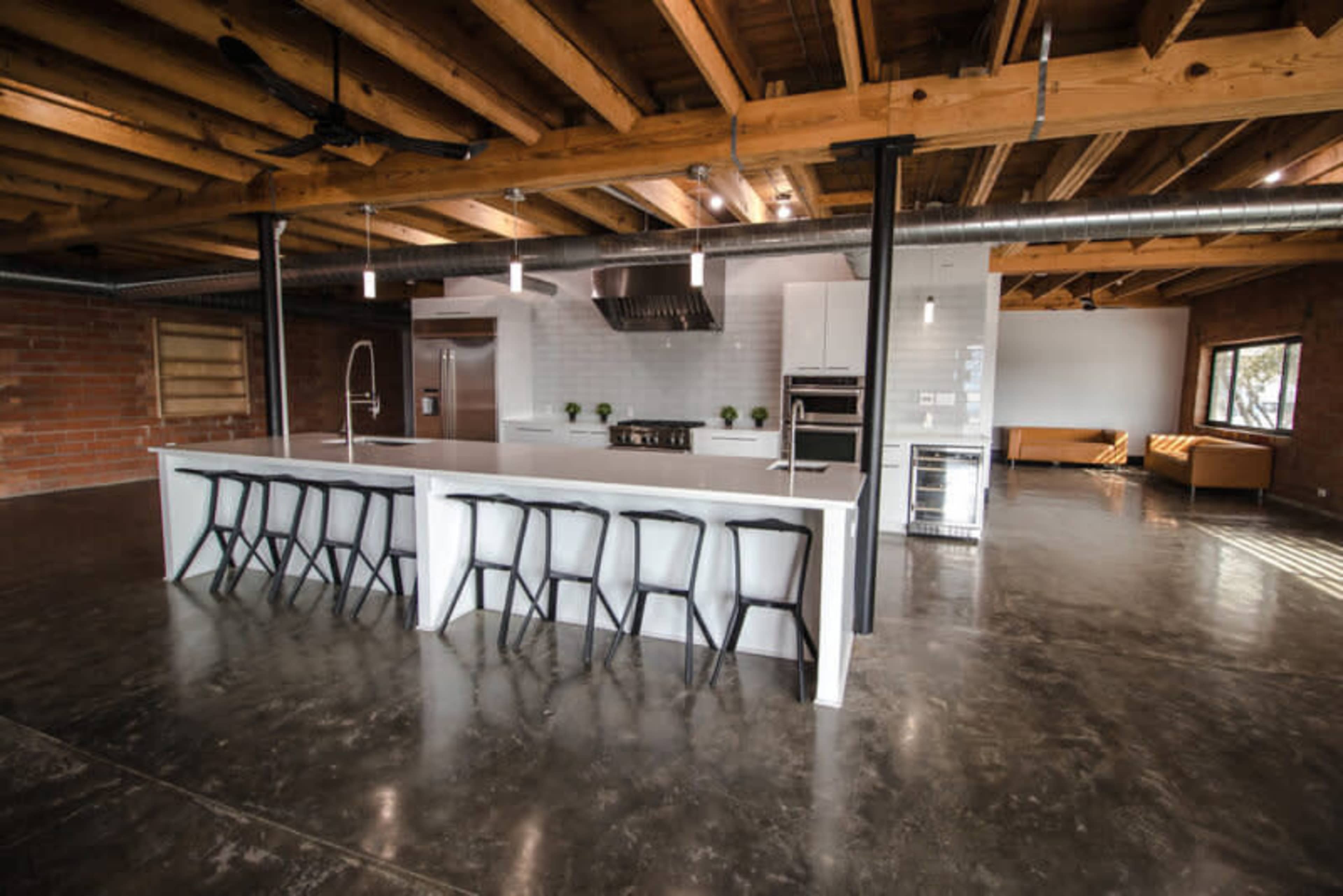 The image shows a spacious modern kitchen with a large white island, eight black bar stools, stainless steel appliances, and exposed wooden beams.