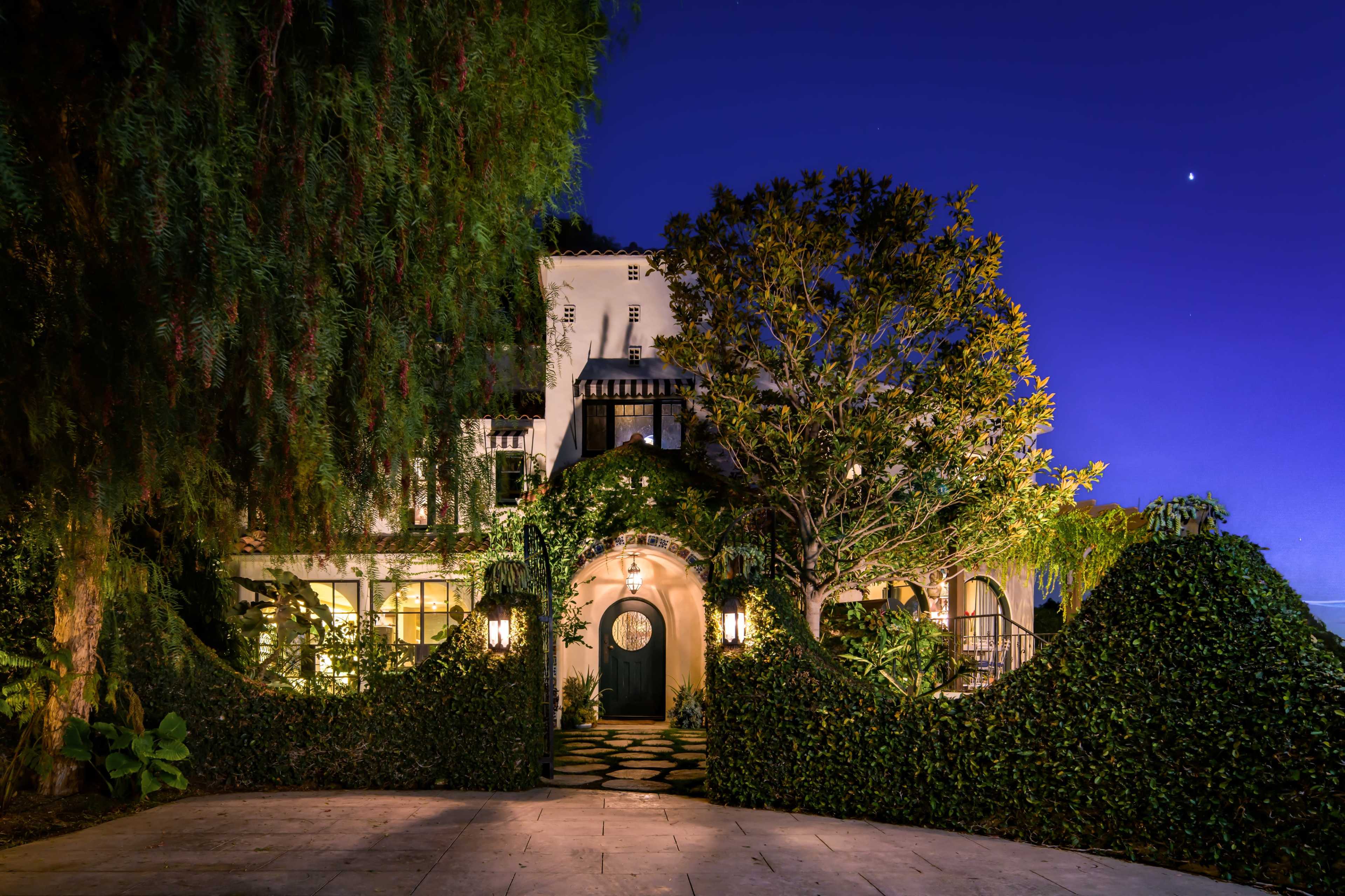 A well-lit, ivy-covered house with a circular front door is surrounded by lush greenery under a twilight sky.