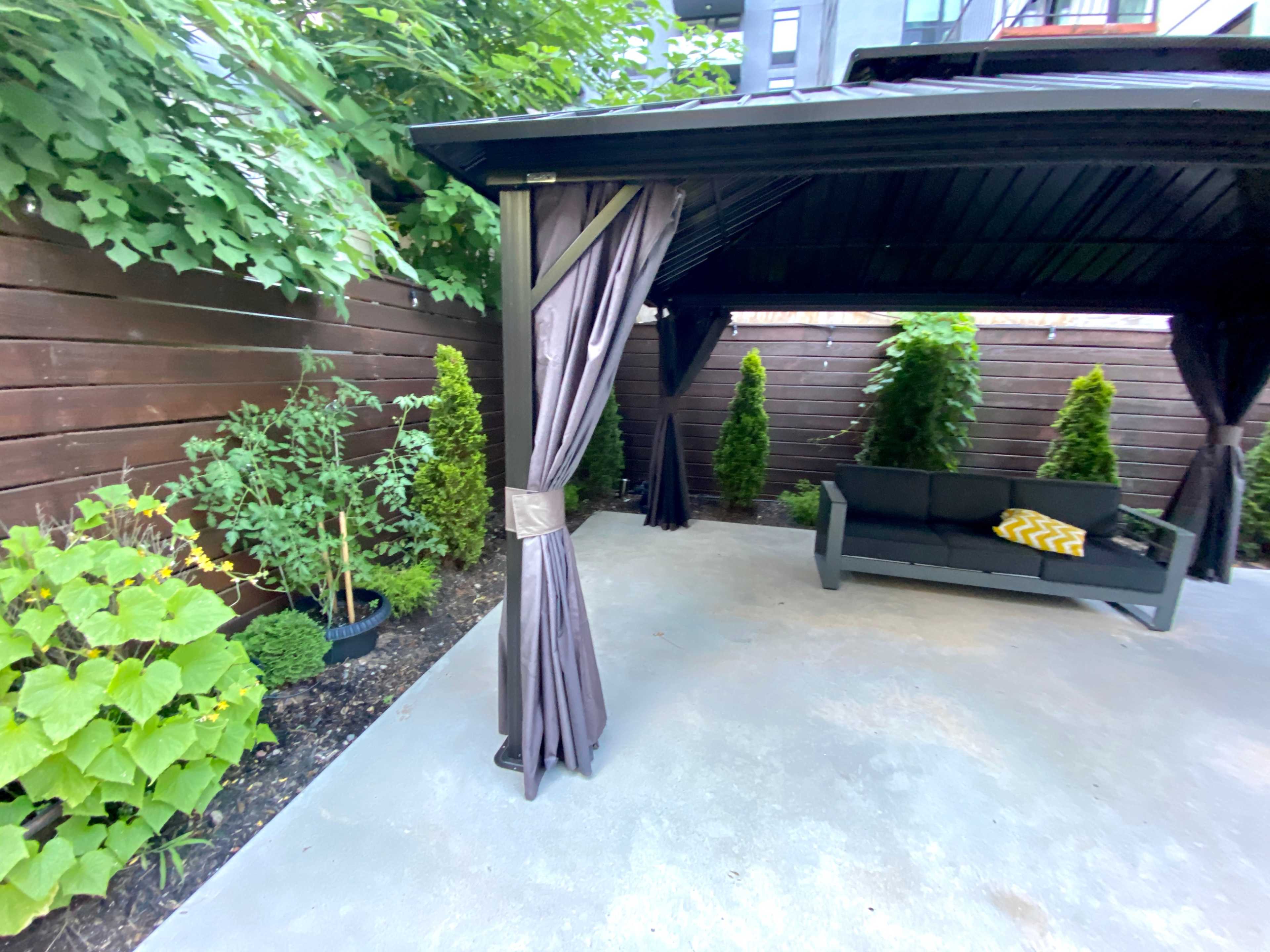 A covered outdoor seating area features a gray sofa and potted plants surrounded by a wooden fence and greenery.