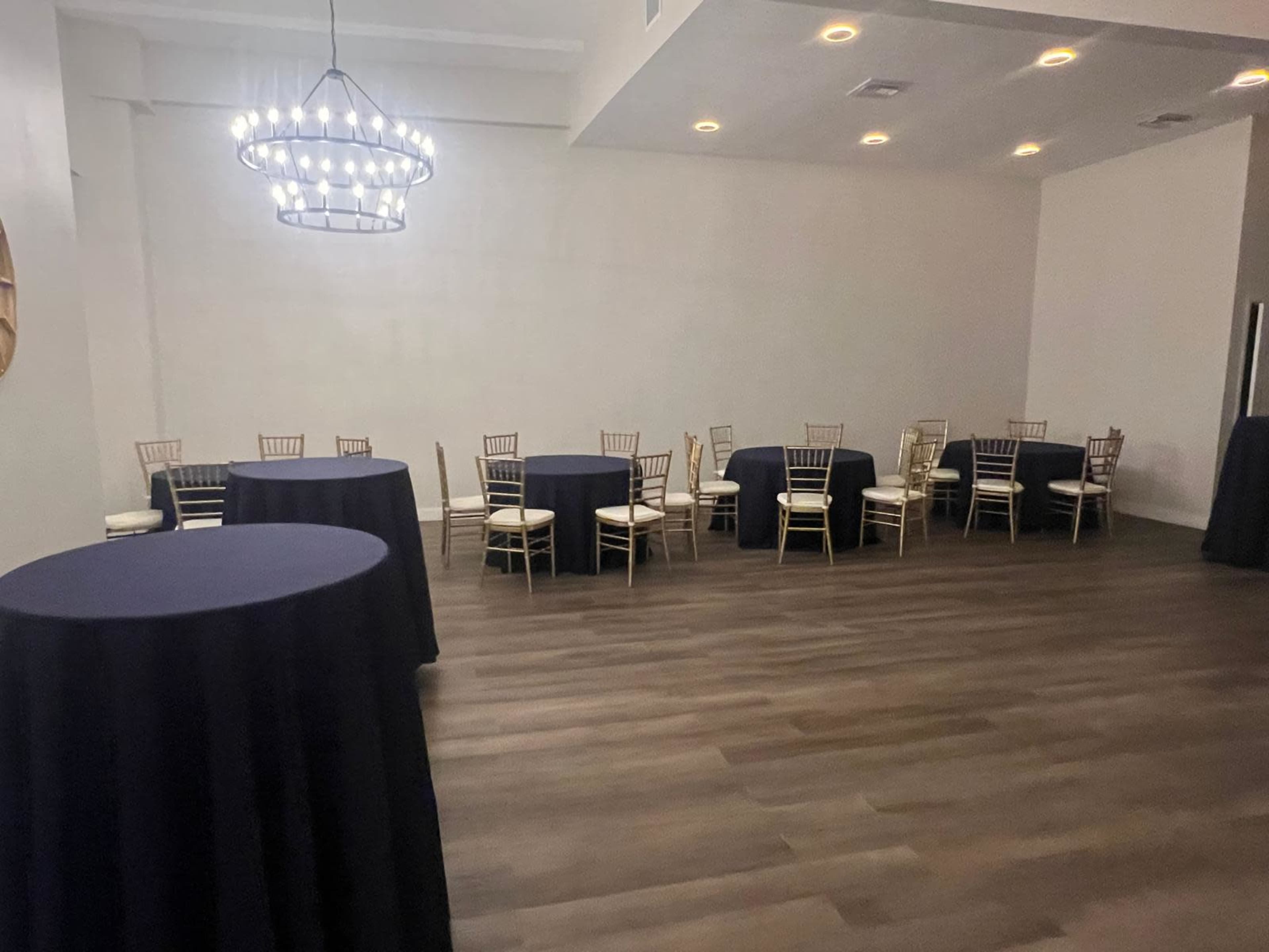 The image shows a spacious interior room with several round tables covered in dark cloths and gold chairs arranged around them, under a modern chandelier.