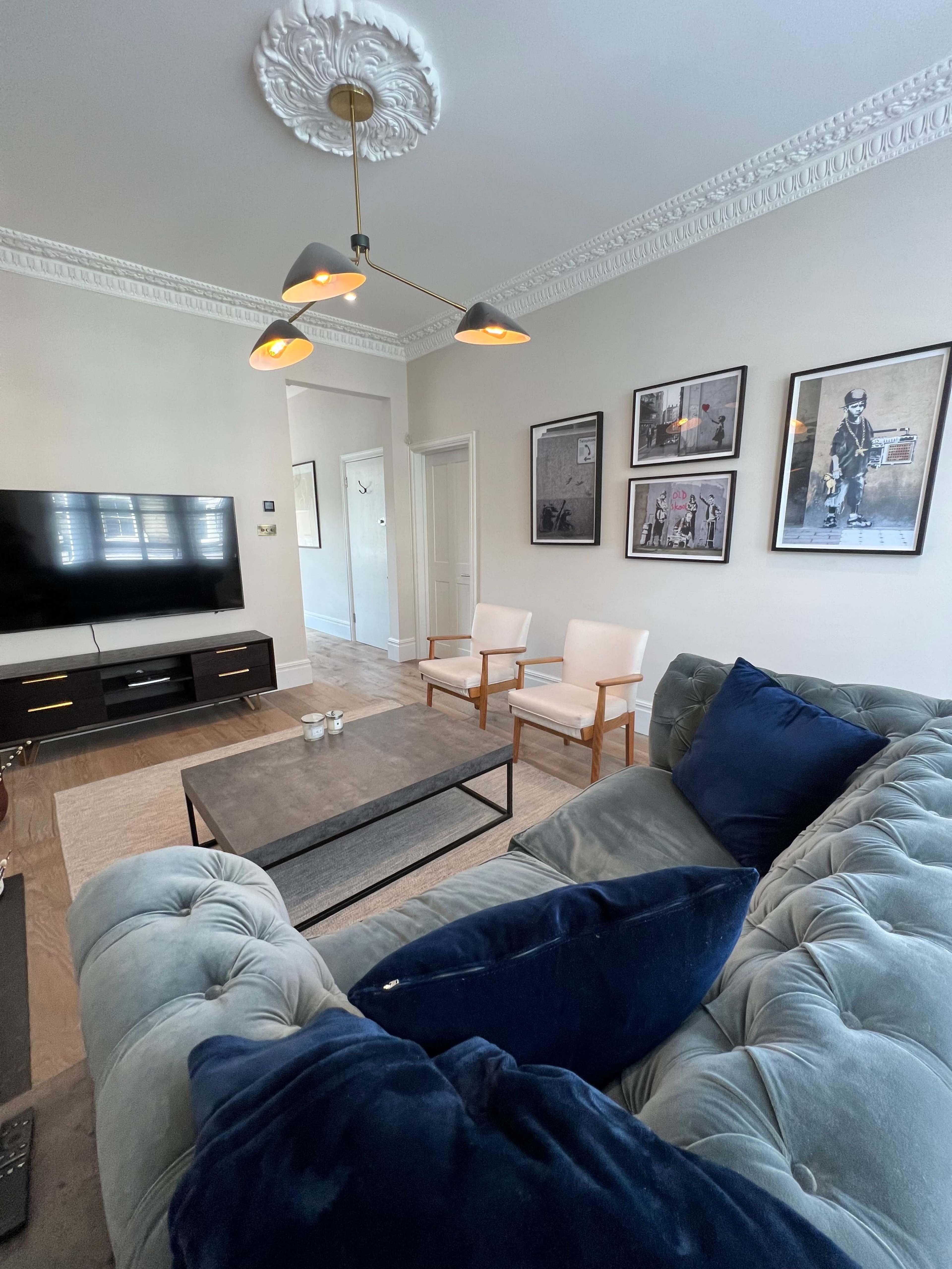 The image shows a modern living room with a gray tufted sofa, a coffee table, two armchairs, a television on the wall, and framed artwork.