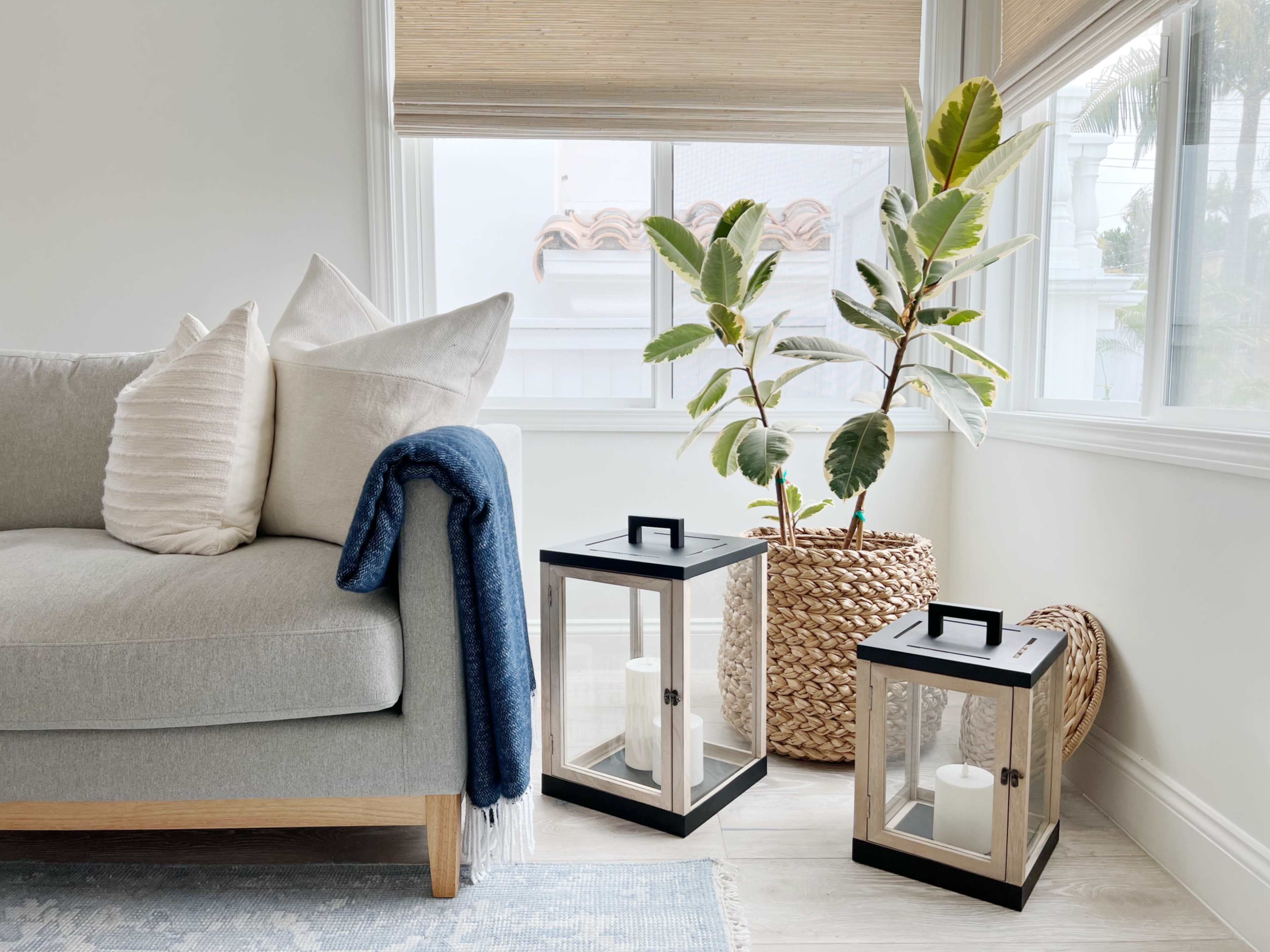 A cozy living room corner features a gray sofa with decorative pillows, a green potted plant, and two lanterns on the floor beside a woven basket.