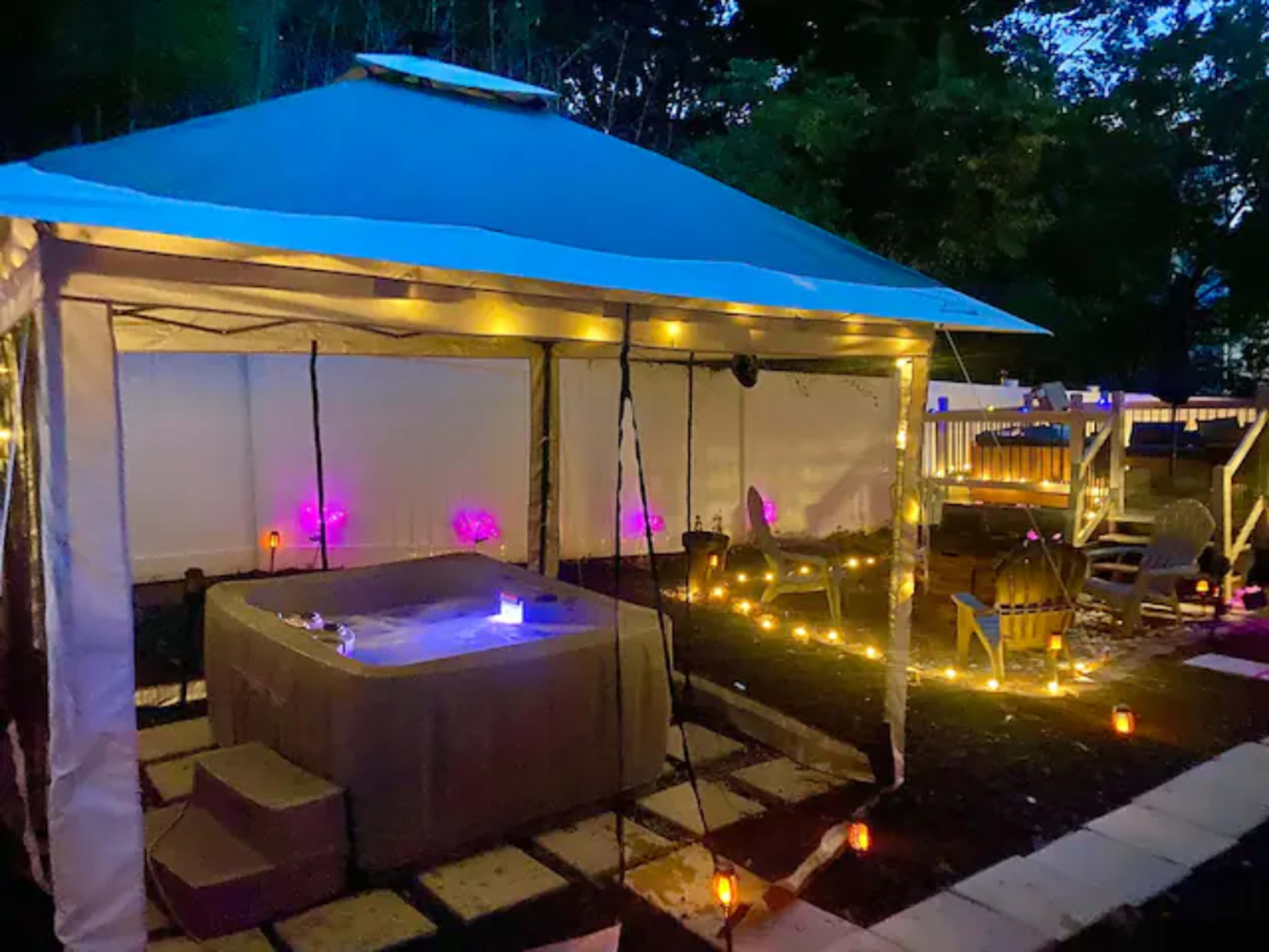 A gazebo with soft lighting surrounds a hot tub in a landscaped outdoor area at dusk.