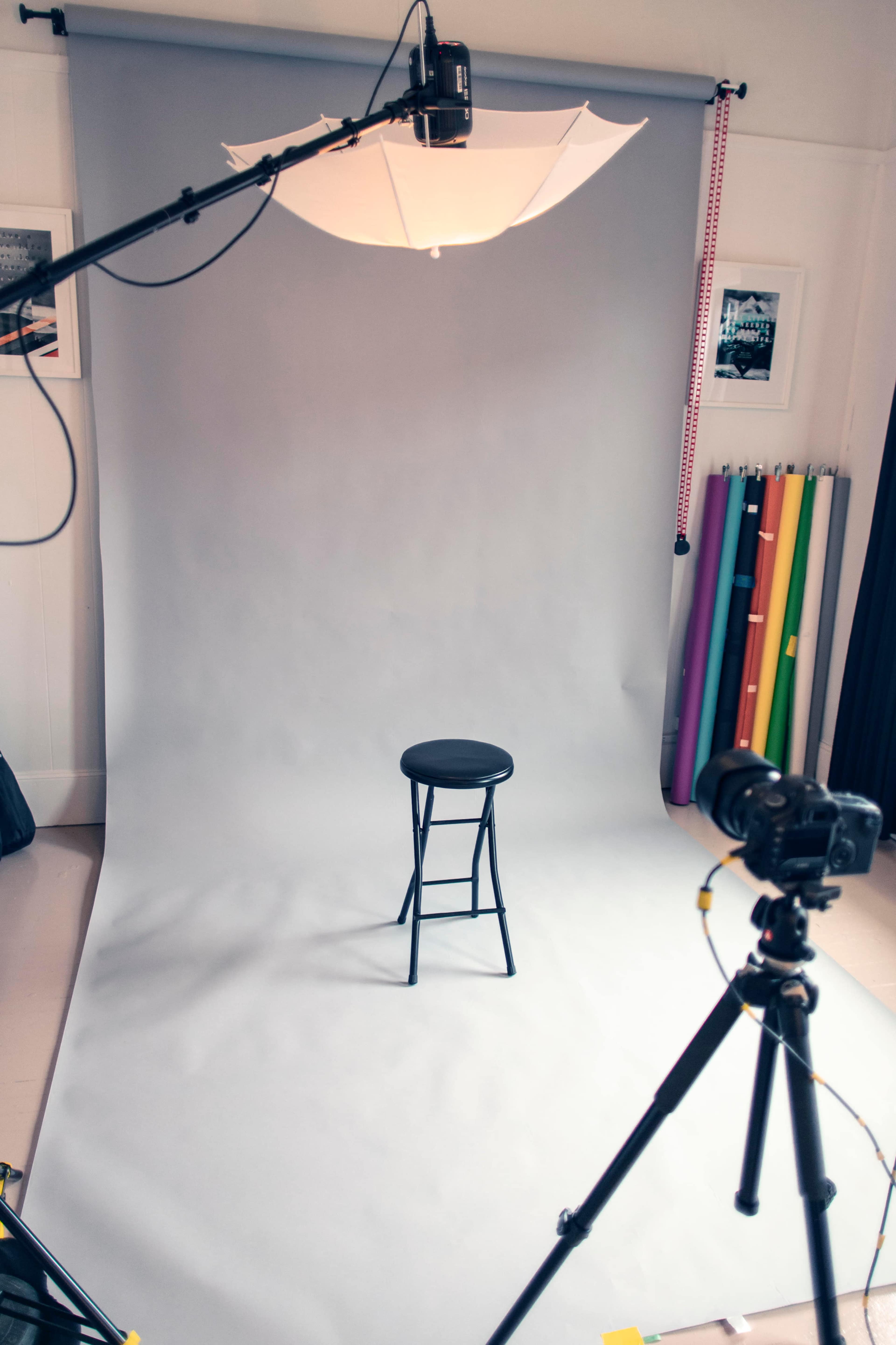 The image shows a photography studio setup with a gray backdrop, a black stool in the center, and camera equipment positioned around it.