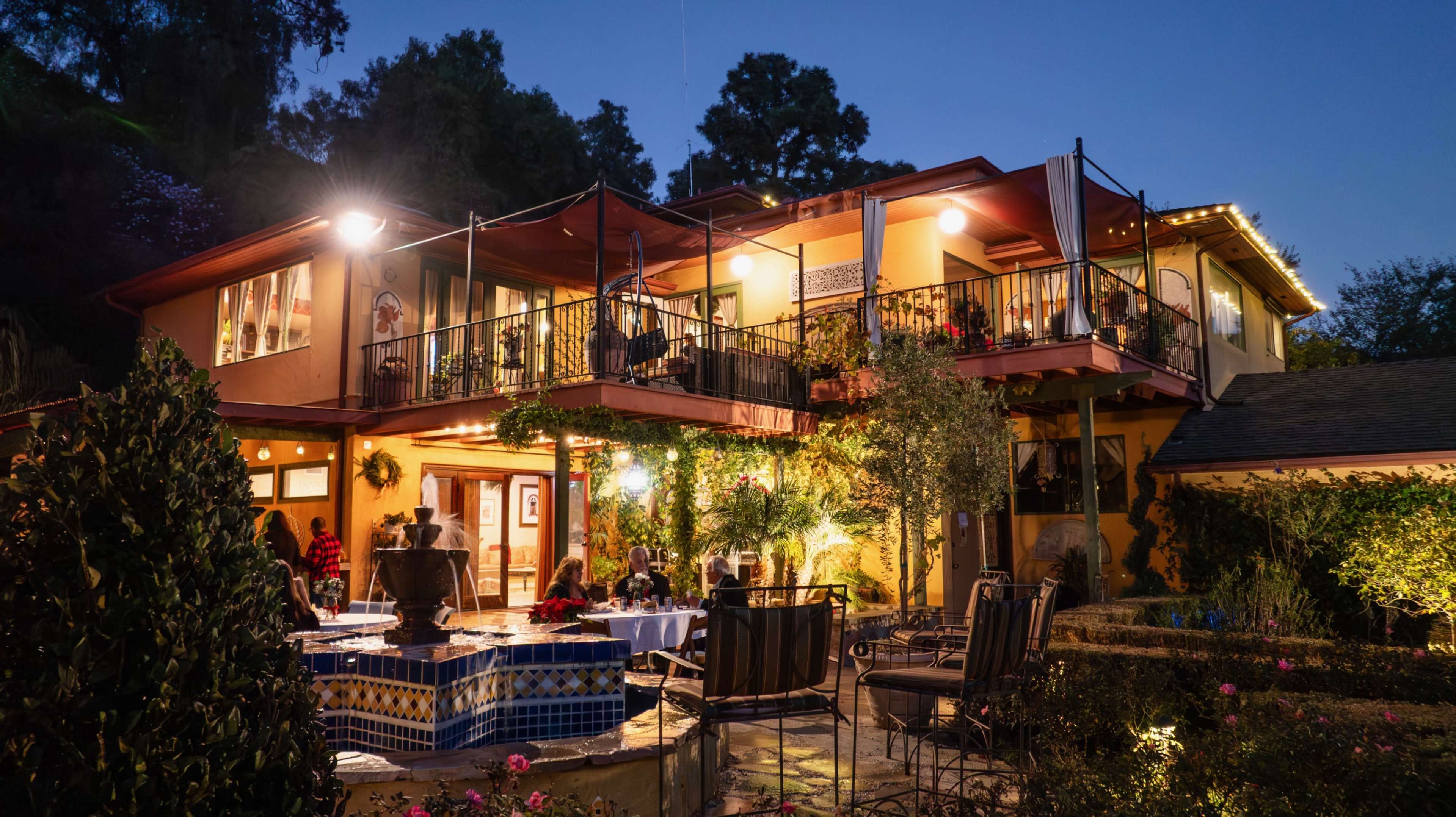 A two-story house with outdoor seating and decorative lighting is set in a garden at dusk.