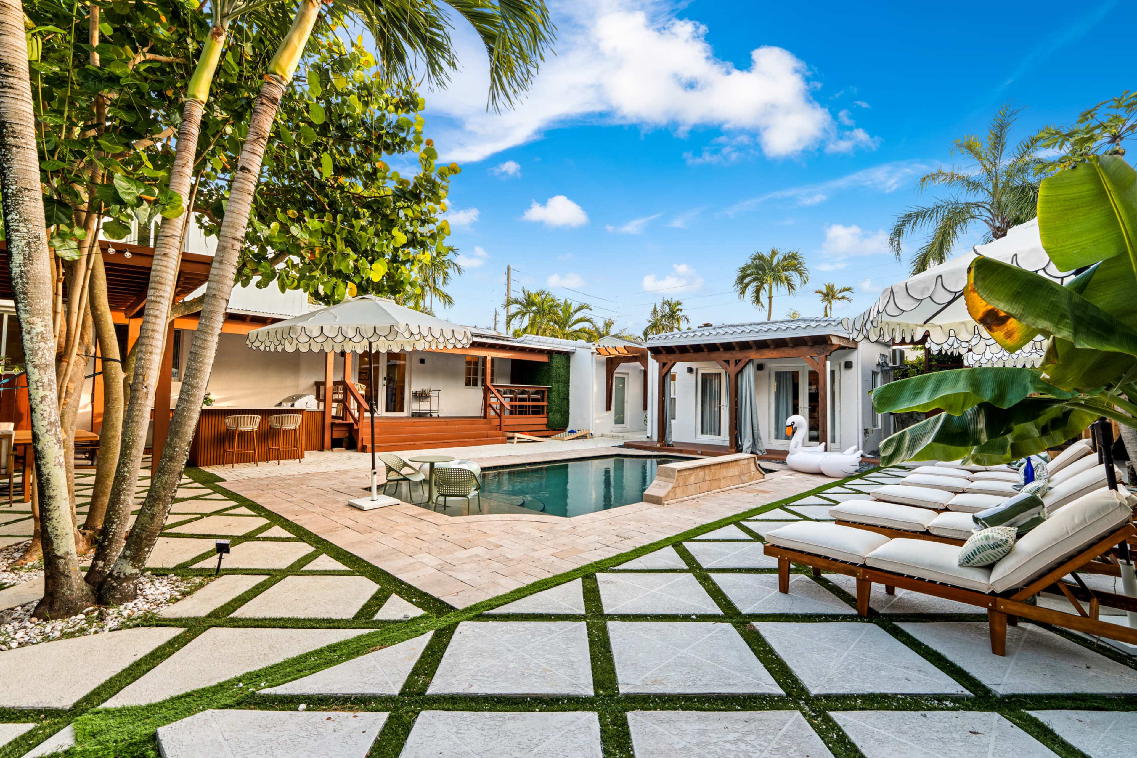 The image features a landscaped outdoor area with a swimming pool, sun loungers, and shaded umbrellas surrounded by palm trees and decorative stone pathways.