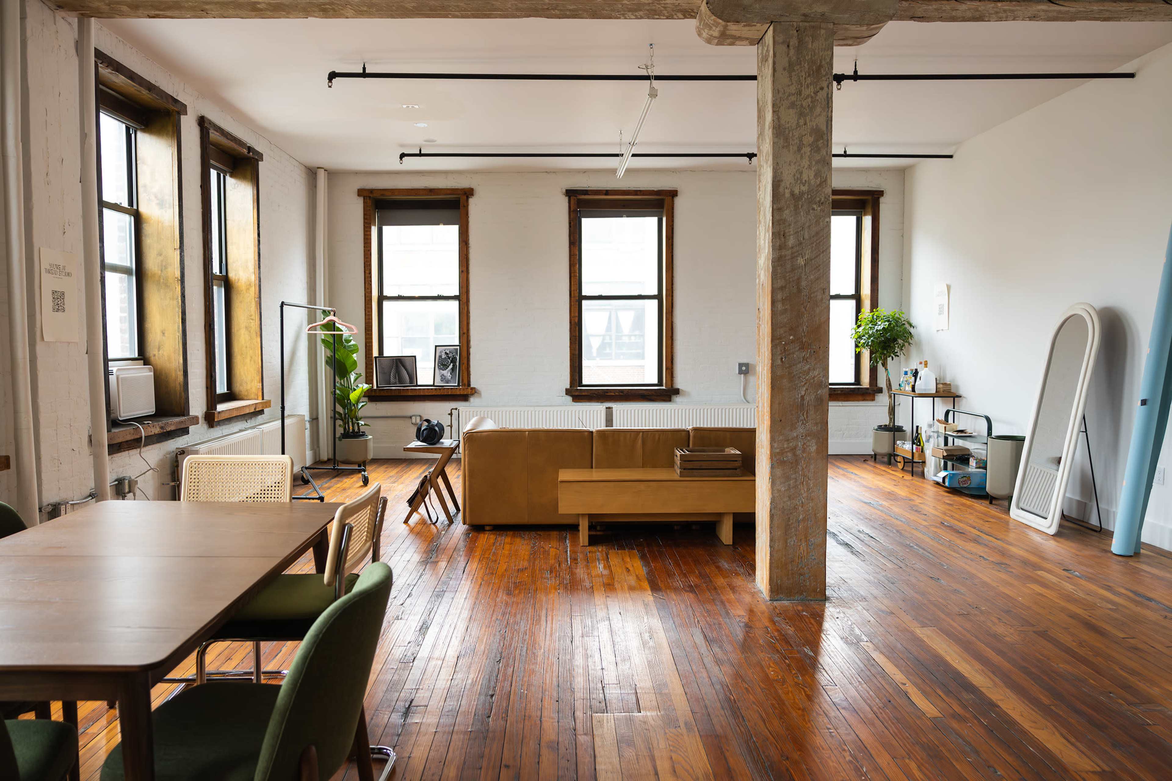 A spacious loft-style living area with large windows, wooden floors, and a mix of furniture including a sofa, dining table, and plants.