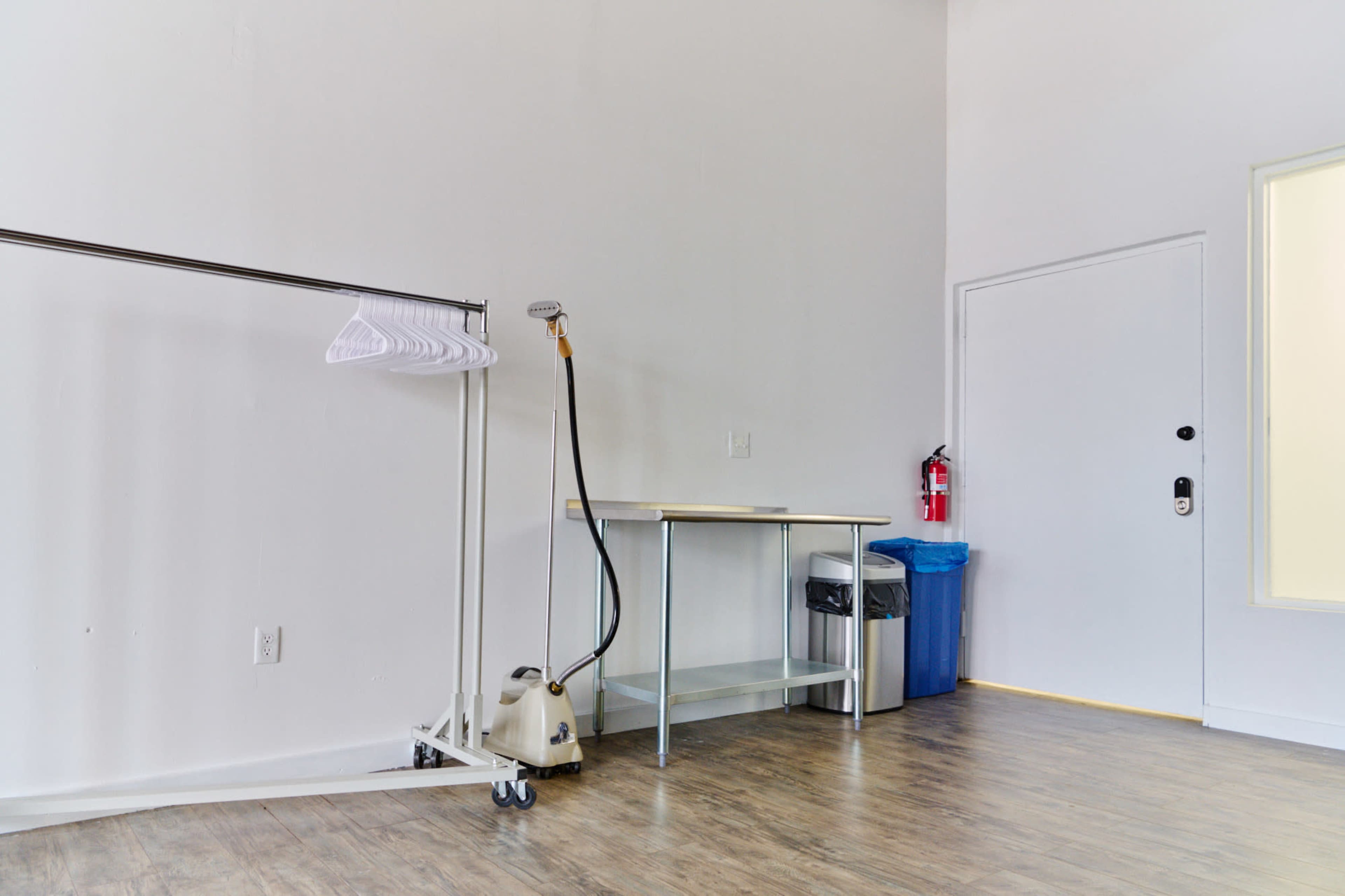 A bare room with a clothing rack, a steam cleaner, a table, a trash can, and a door.