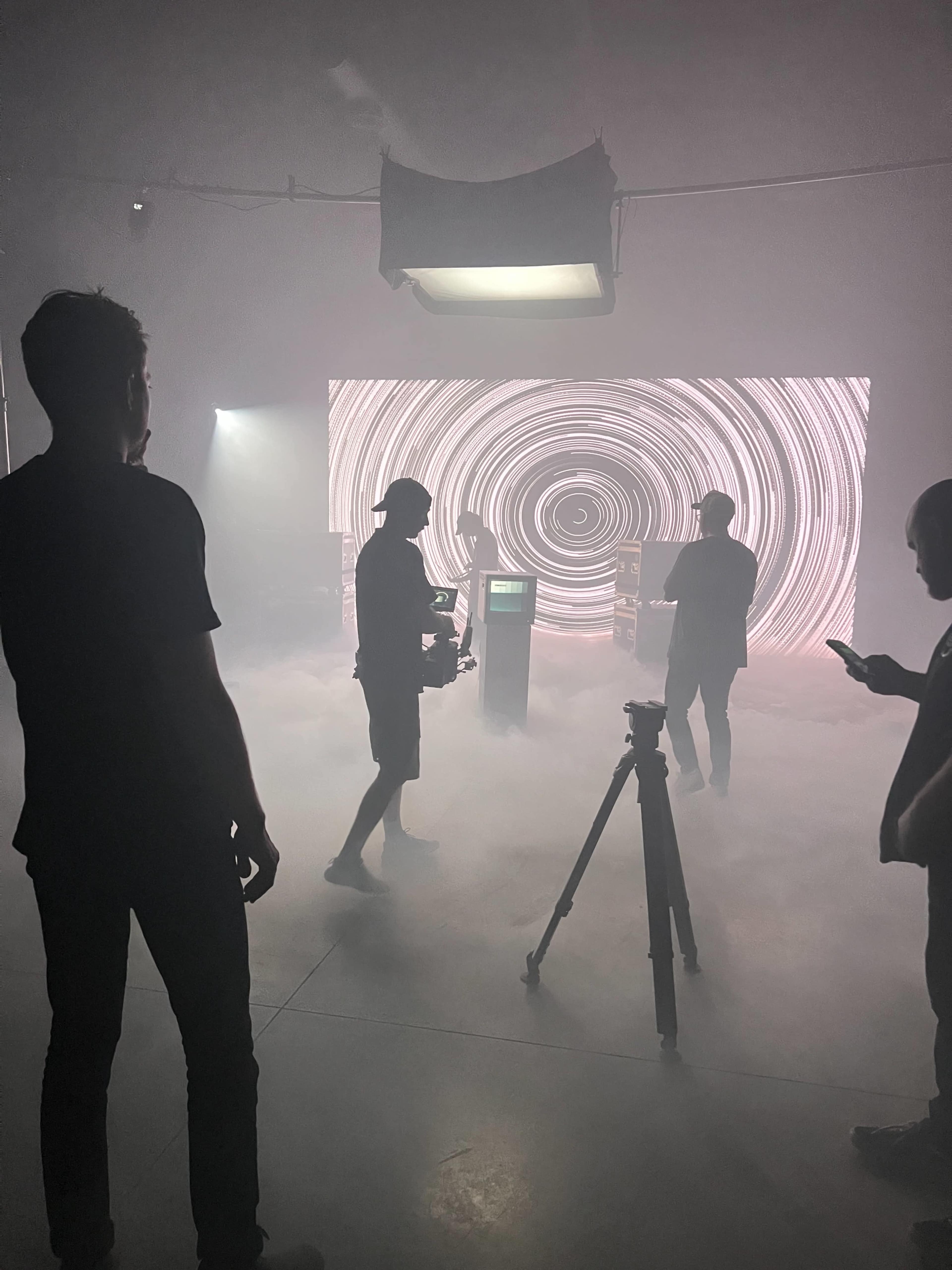 A group of individuals sets up a scene in a fog-filled space with a swirling light display in the background.